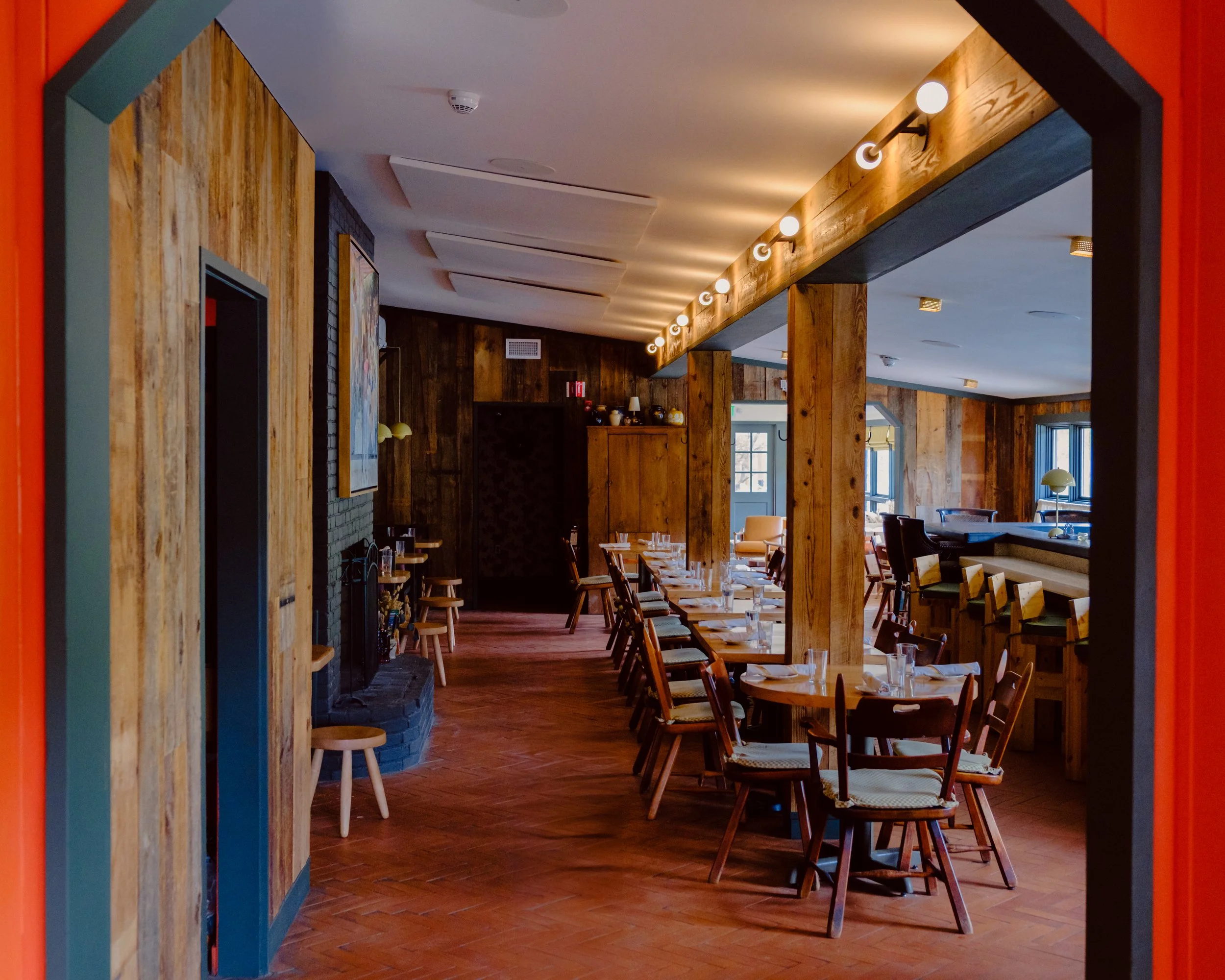 Interior of a restaurant with wooden walls, tables set with glasses and napkins, and chairs around the tables. The ceiling has decorative elements and lighting fixtures, and there are windows letting in natural light.
