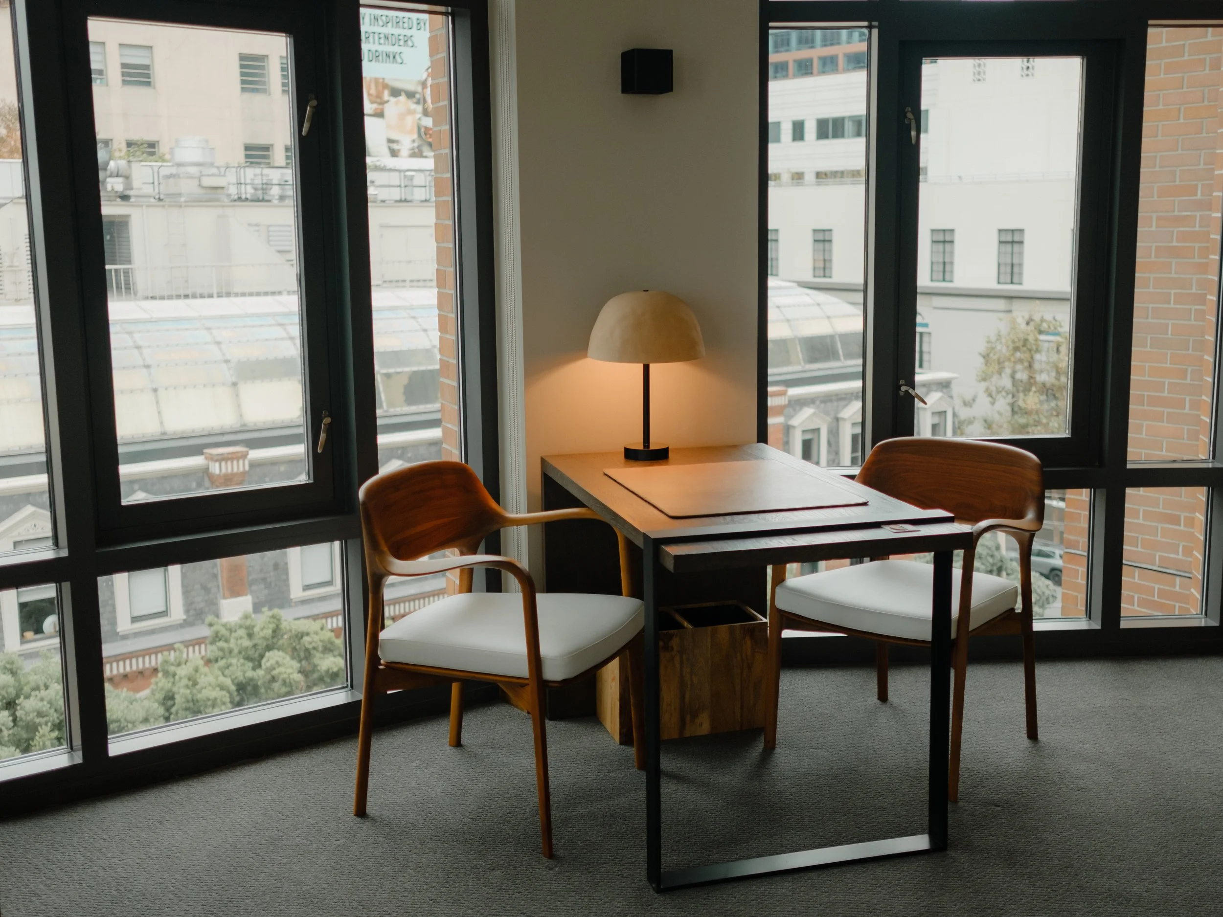 A cozy interior space with reading chairs around a table, near windows showing a cityscape with buildings and trees outside, and a standing lamp providing warm light.