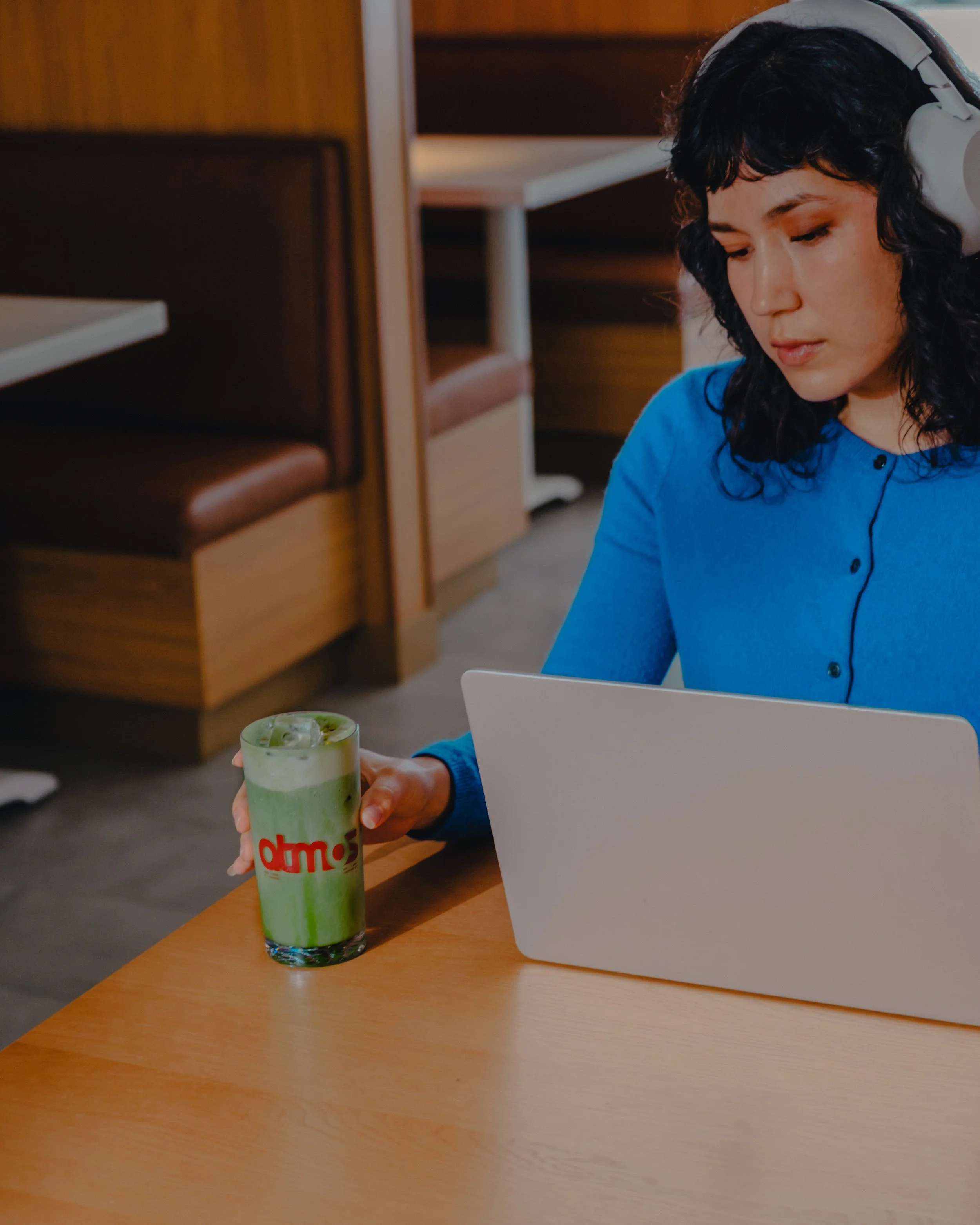 A woman with dark curly hair wearing a blue shirt and white headphones sits at a wooden table with a laptop. She is holding a tall glass with a green beverage that has whipped cream on top. In the background, there are wooden chairs and tables in a c