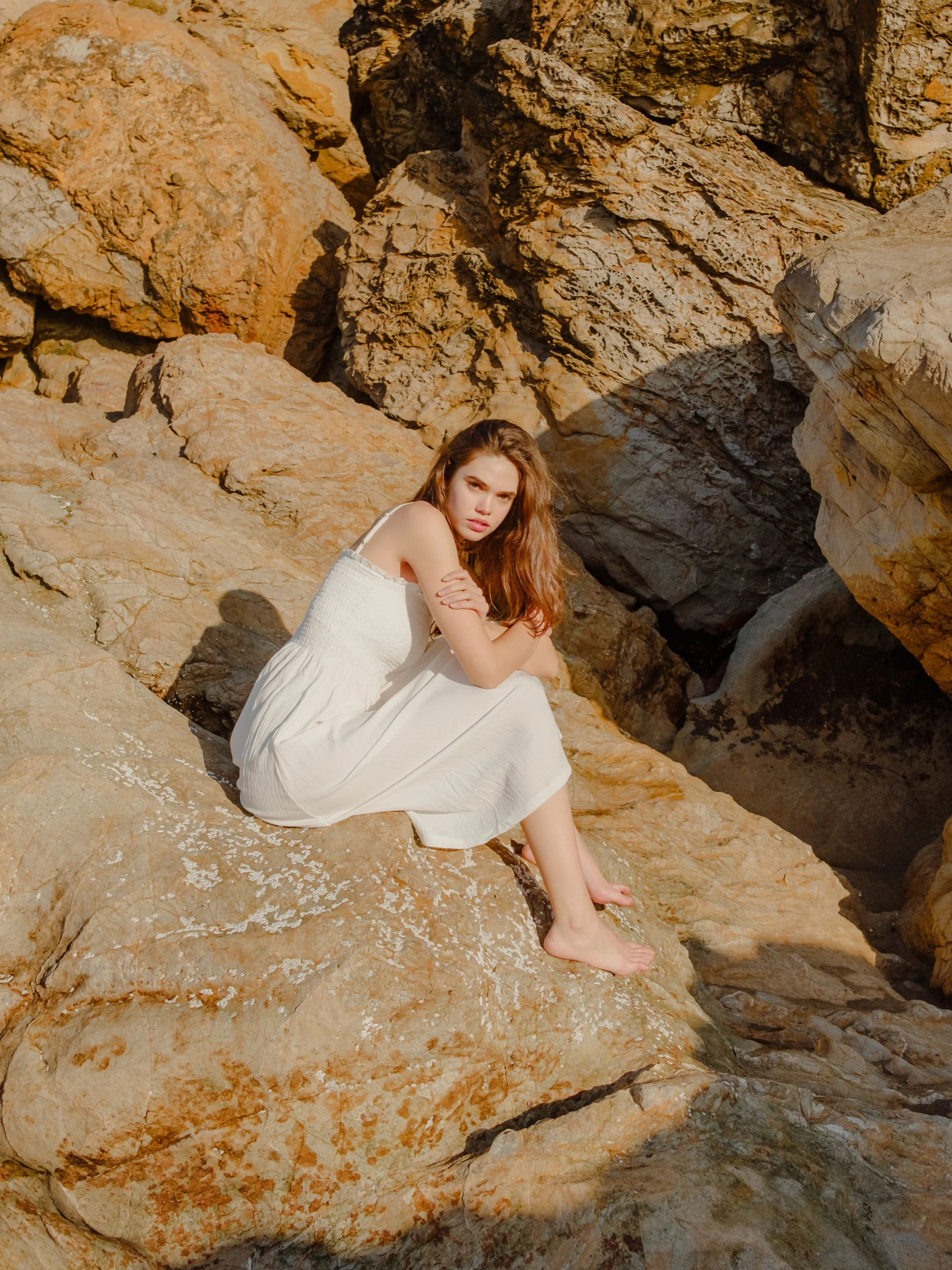 A young woman with long brown hair wearing a white dress sitting on a rocky terrain with large weathered rocks in the background.