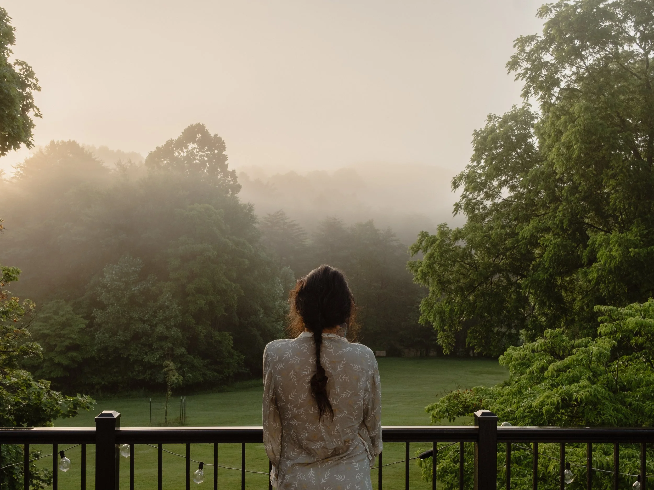 A woman with long brown hair styled in a braid, wearing a light-colored dress with a leaf pattern, stands on a deck overlooking a foggy, lush green landscape with trees and a grassy area.