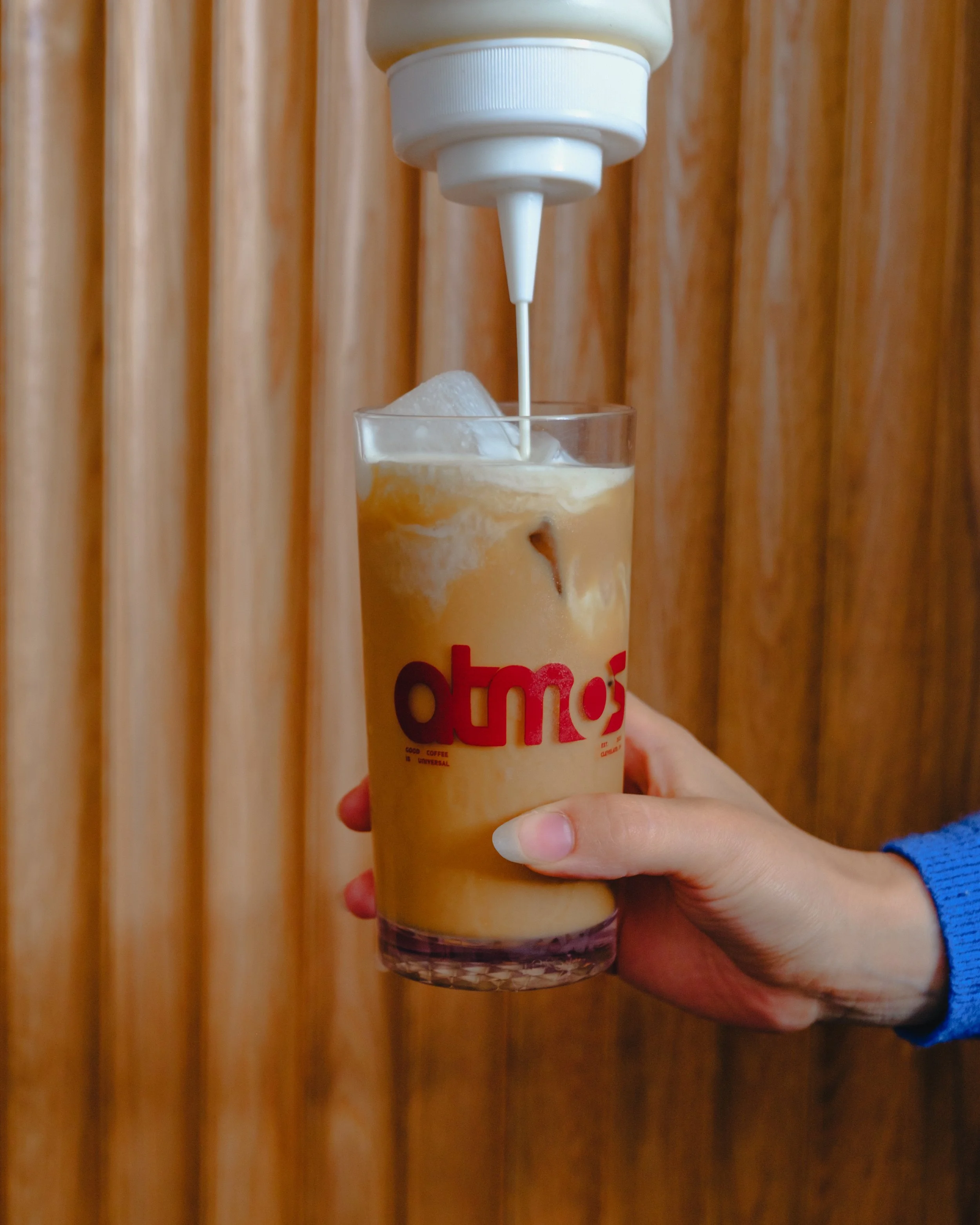 A hand holding a plastic cup filled with iced coffee, with some milk or cream being poured into it from a white squeeze bottle, a wooden background.
