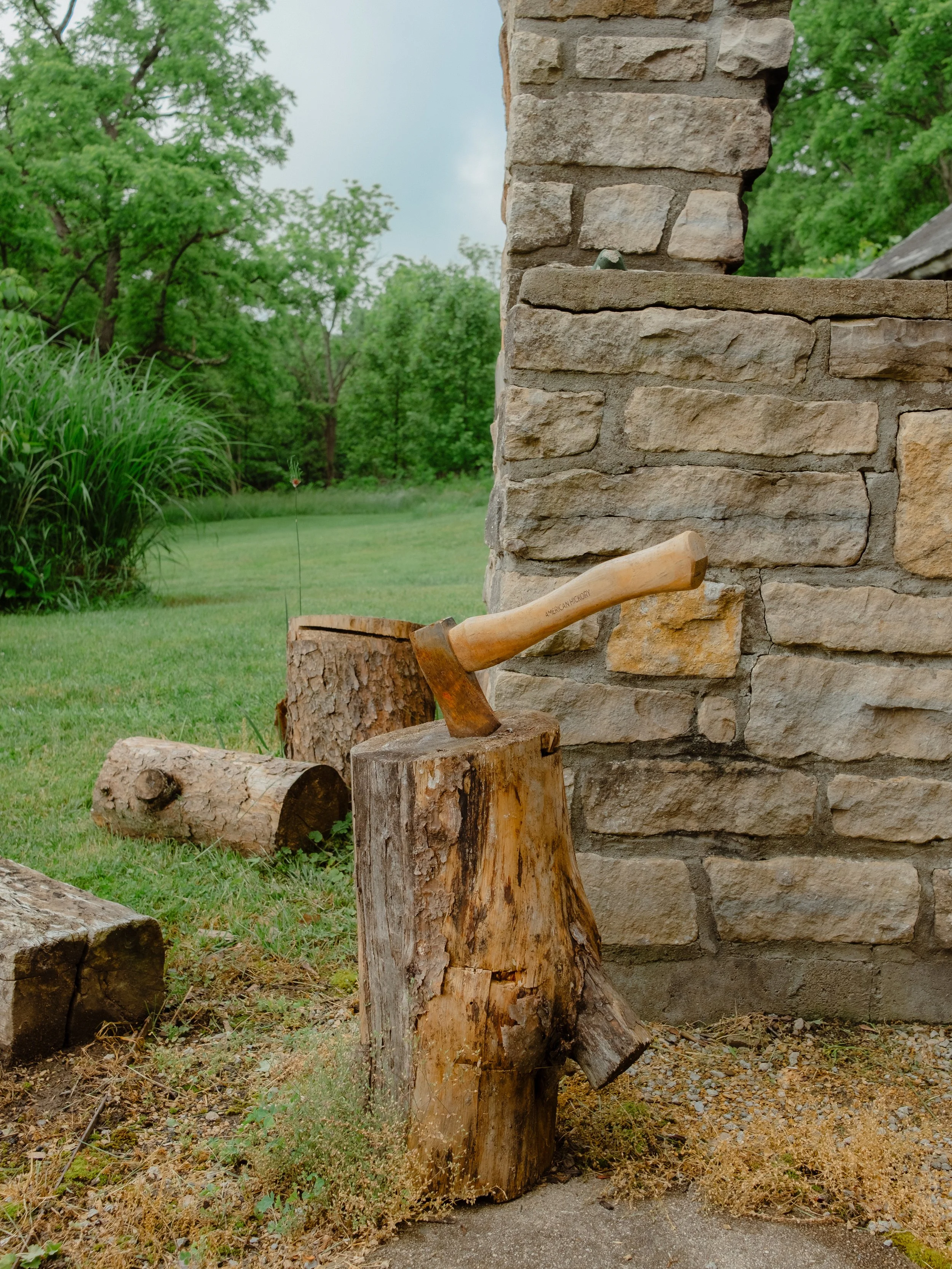 A stone wall with a hatchet stuck in a large piece of wood in front of it, surrounded by grass, weeds, and logs, with green trees in the background.