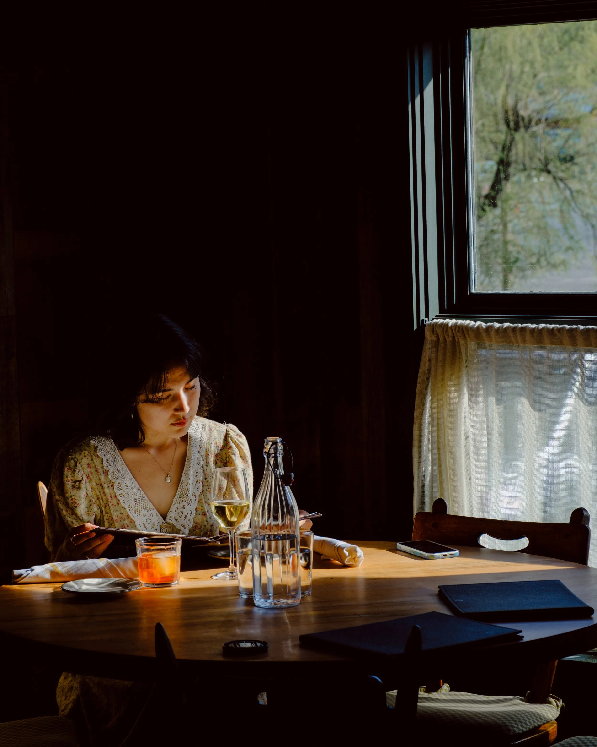 A woman with dark curly hair wearing a floral dress sitting at a wooden table with a glass of white wine, bottles, and a smartphone inside a dimly lit room, with sunlight coming through a window showing trees outside.