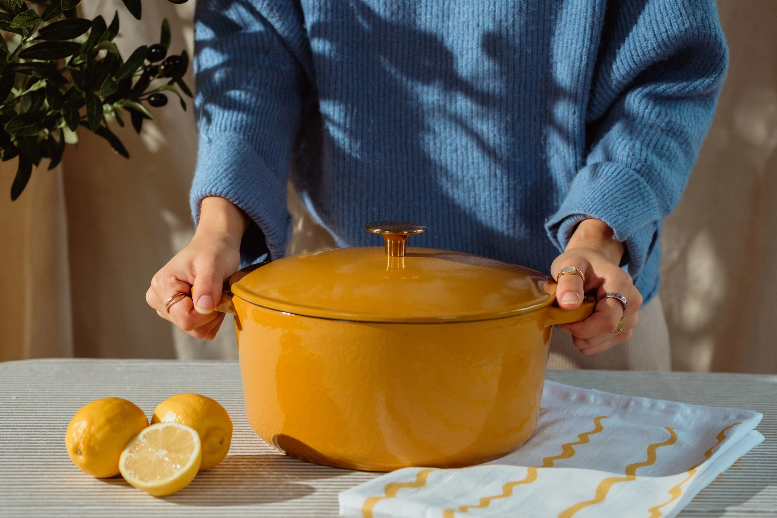 A person wearing a blue sweater holding a yellow pot on a table with lemons around it. There are whole and sliced lemons on the table, with a cloth having yellow and white zigzag pattern. A potted plant is visible on the left side.
