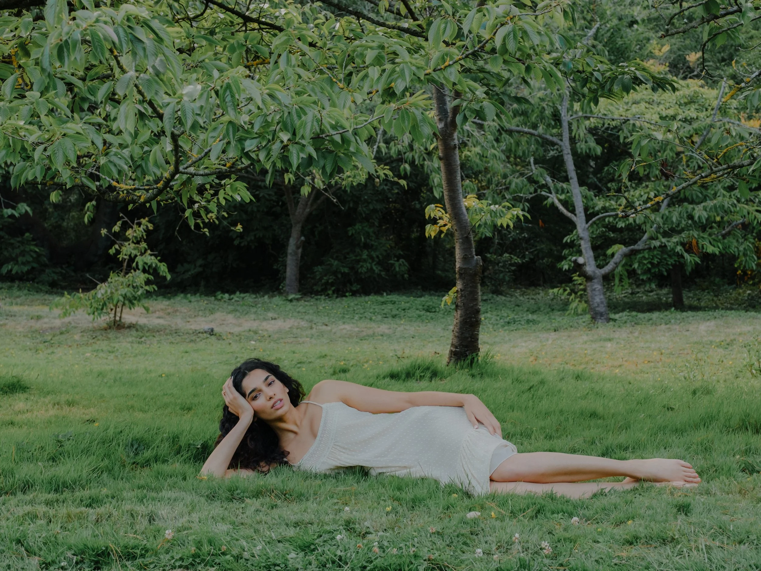 A woman with dark curly hair lying on green grass in a park, with trees in the background. She is wearing a light-colored sleeveless dress and resting her head on her hand, looking at the camera.