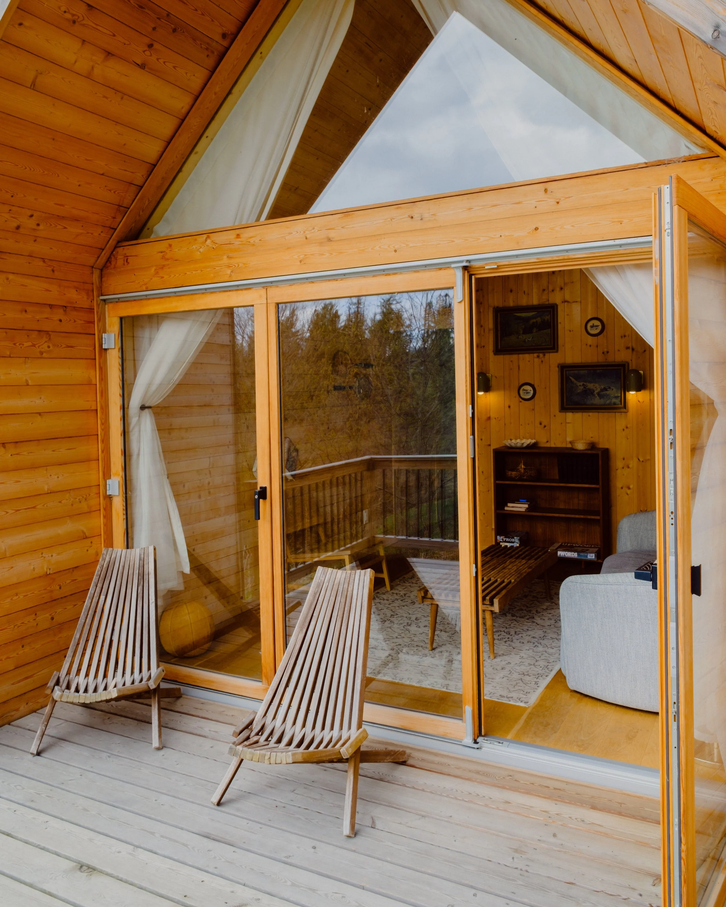 A cozy wooden porch with two wooden chairs placed on a light-colored wooden deck, adjacent to a room with wood-paneled walls and large glass doors leading inside.