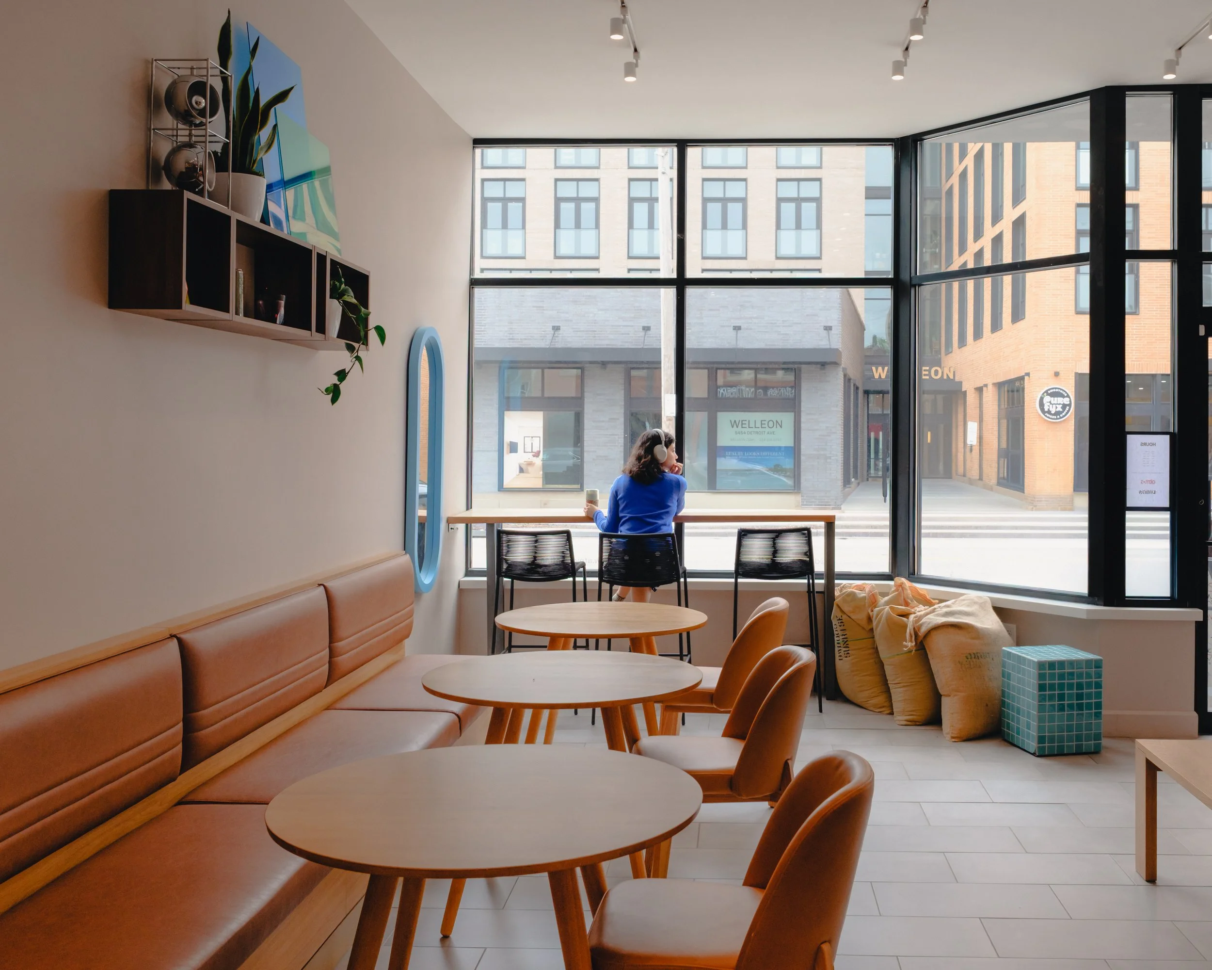 Interior of a modern coffee shop with wooden tables and chairs, a long leather bench along the wall, and large windows overlooking a city street. A woman with headphones is sitting at a table near the window.