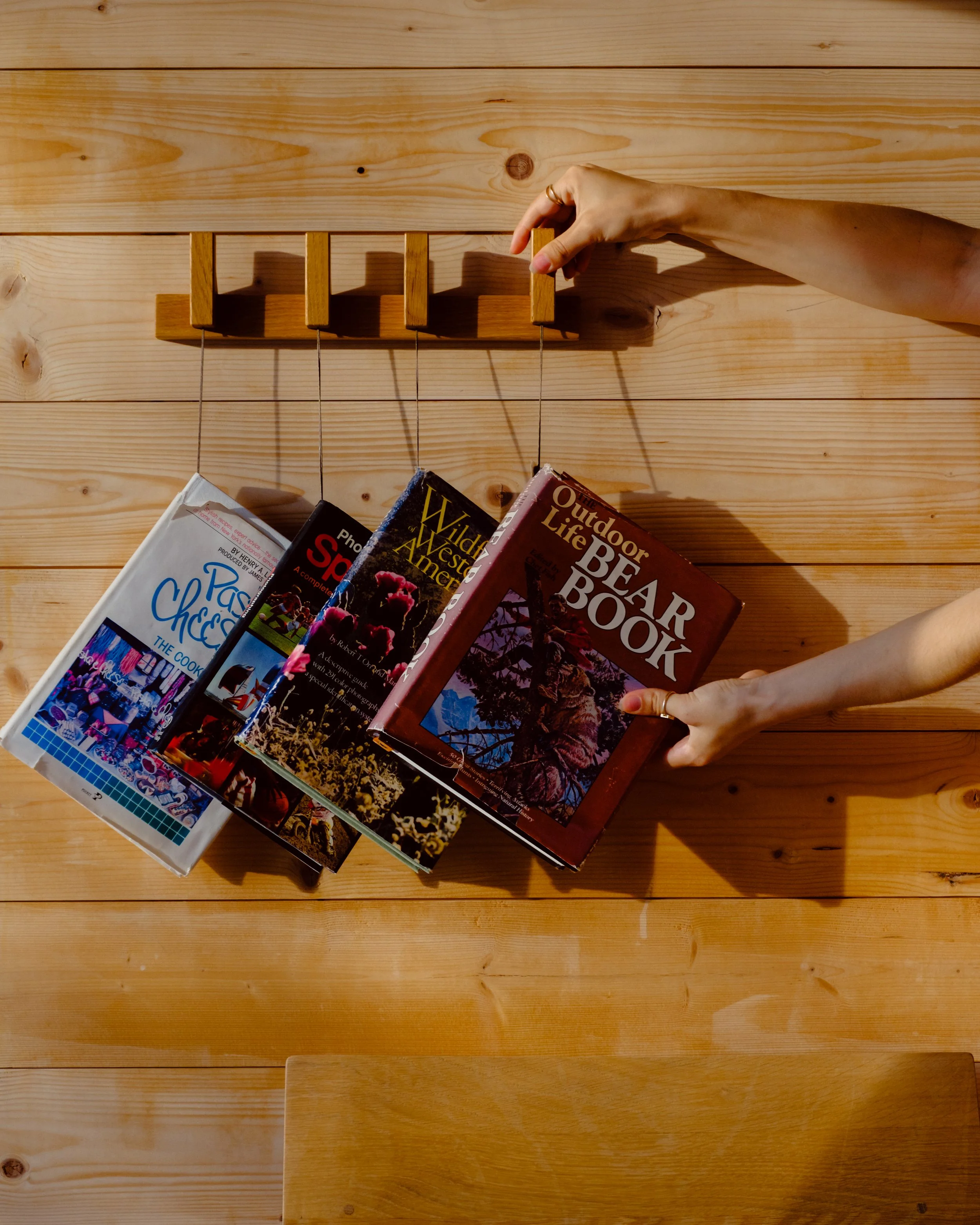 A person's hand adjusting a wooden book rack on a light-colored wooden table. Several books are visible, including titles like 'Classical China,' 'Wild West America,' and 'Outdoor Life Beer Book.'