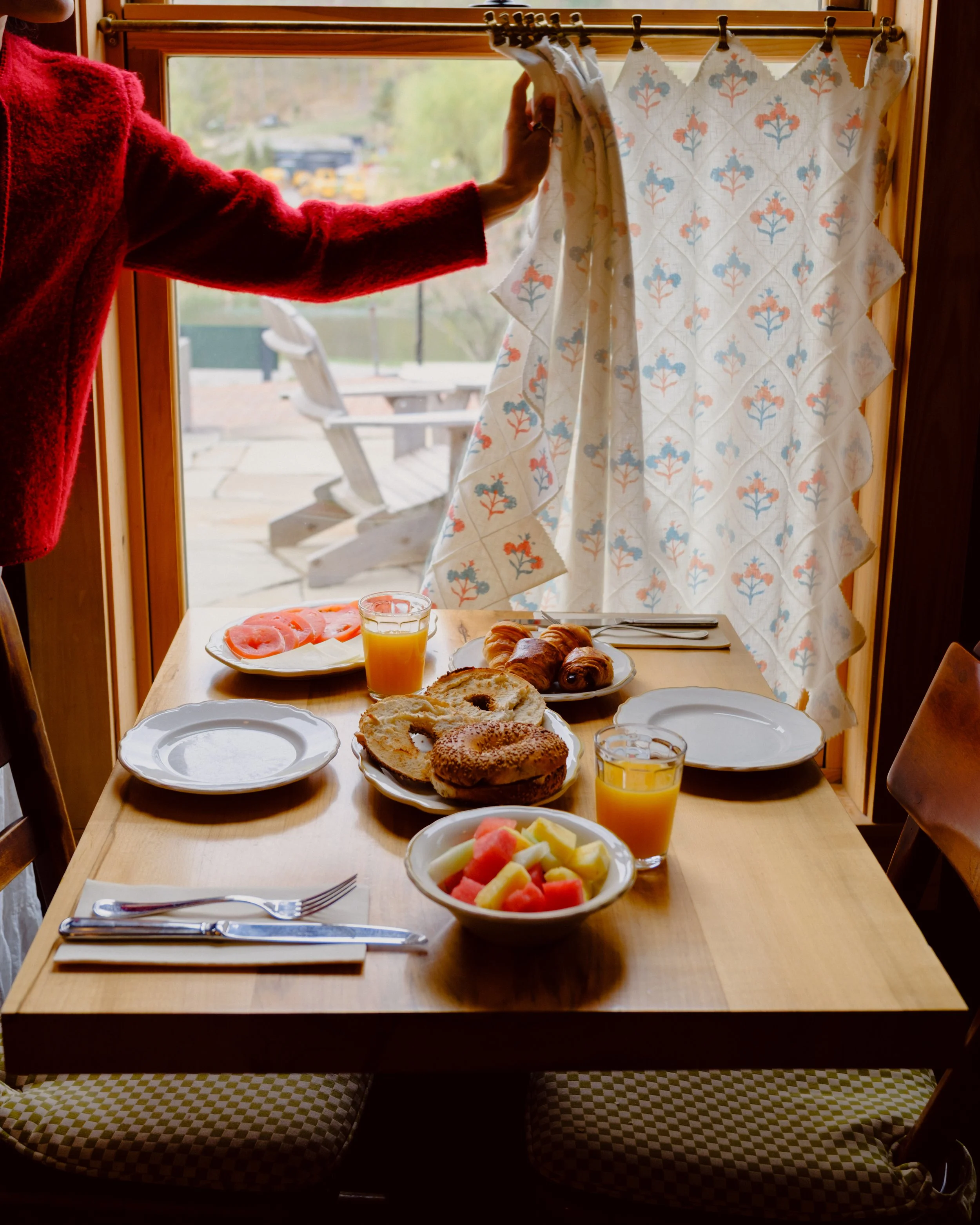 A breakfast table set for four, with plates of sliced tomatoes, a bowl of fruit, croissants, and bagels, along with glasses of orange juice. There's a window with a view of trees and outdoor furniture, and a person in a red sweater is holding back a 