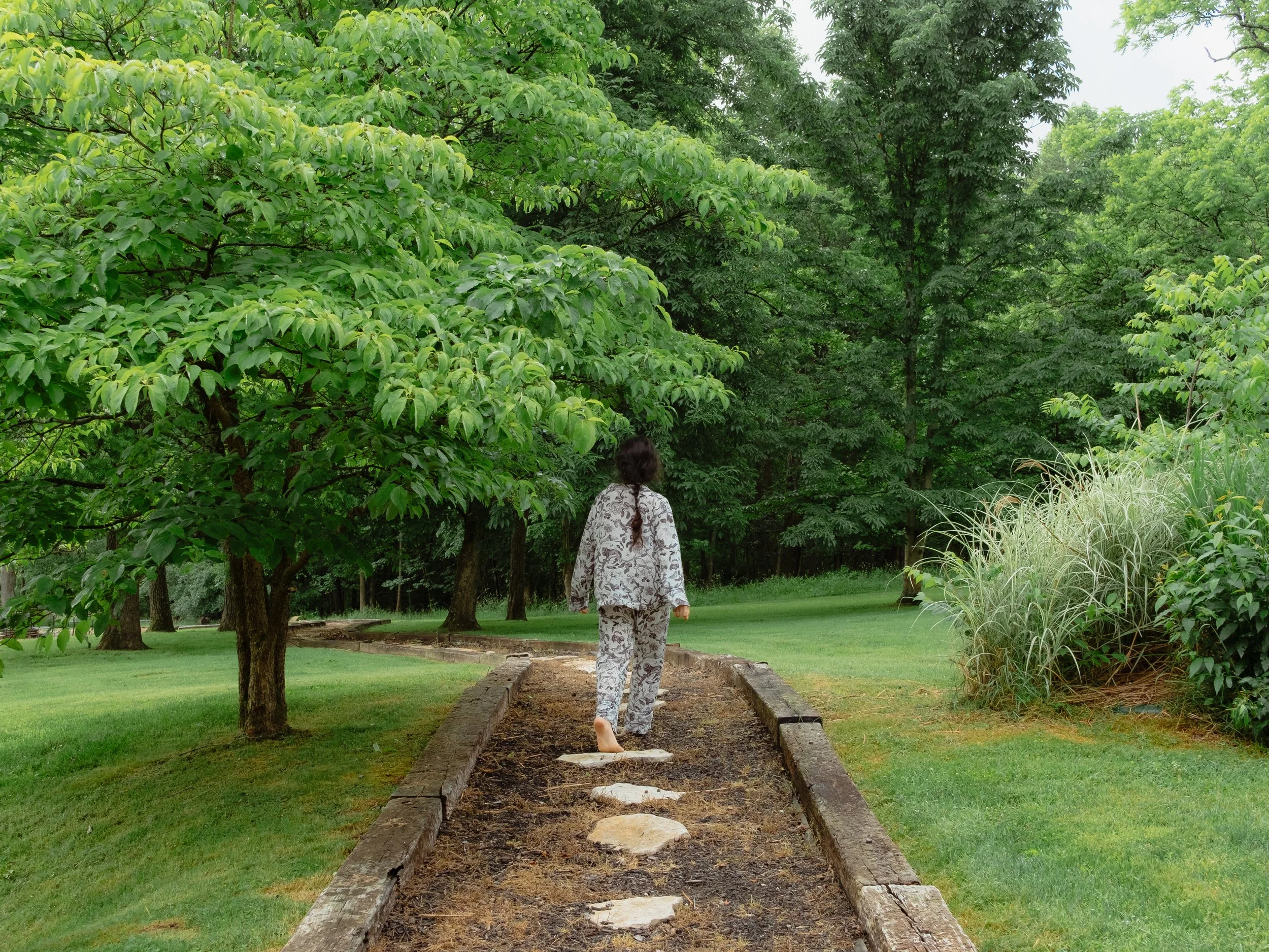 A person with long dark hair in a braid wearing pajamas with a floral pattern walking barefoot on a narrow dirt path bordered by wooden planks through a lush green park with trees and grass.