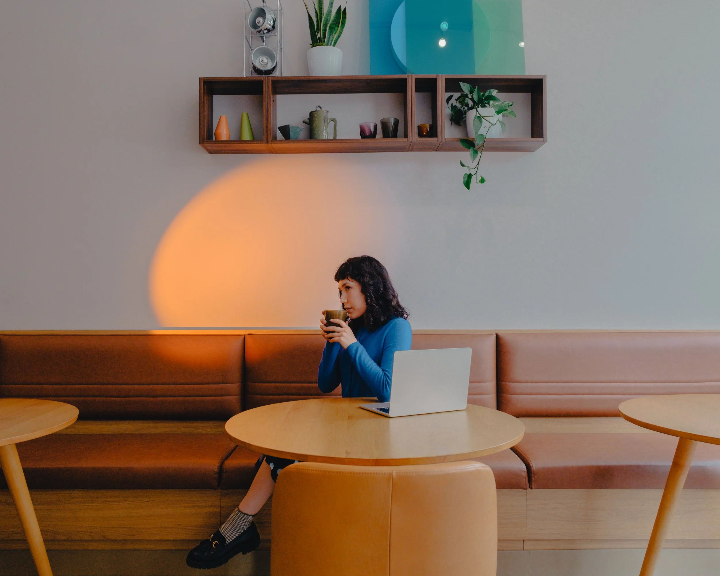 A woman sitting at a wooden table in a cafe holding a cup, with a laptop in front of her. Behind her, there's a long wooden bench with a beige cushion, and a wall shelf with decorative vases, a teapot, and potted plants. The wall is painted light gra