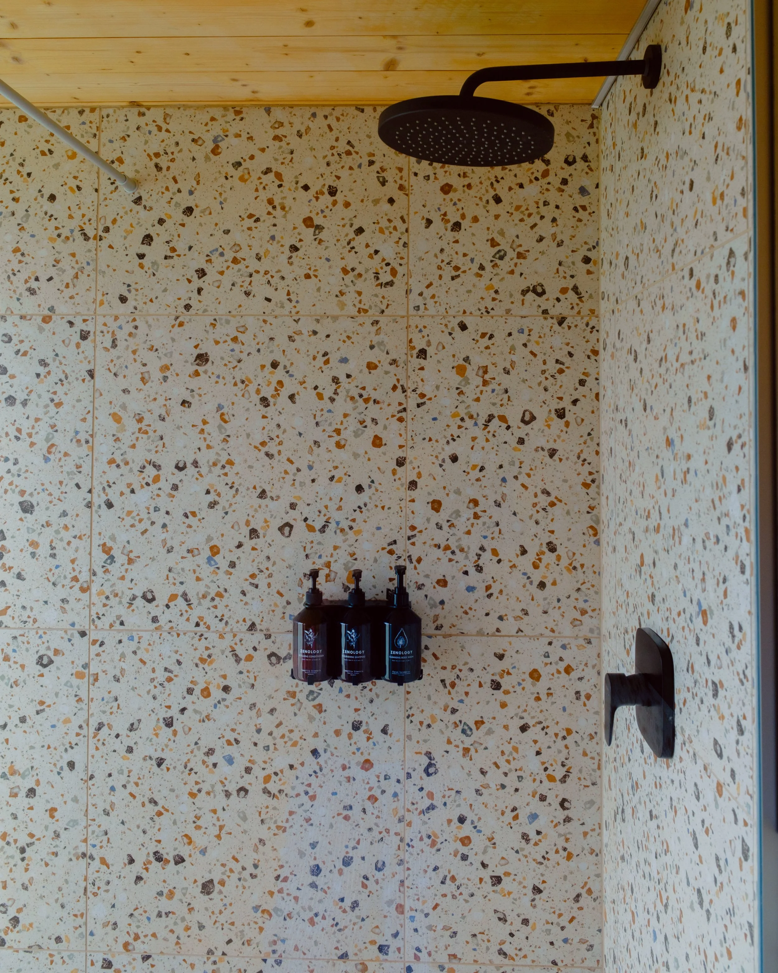 A shower with a black rain showerhead mounted on a tiled wall. Below the showerhead, three black bottles of toiletries are placed on the tiled surface. The shower wall has a terrazzo pattern with multicolored chips on a cream background.