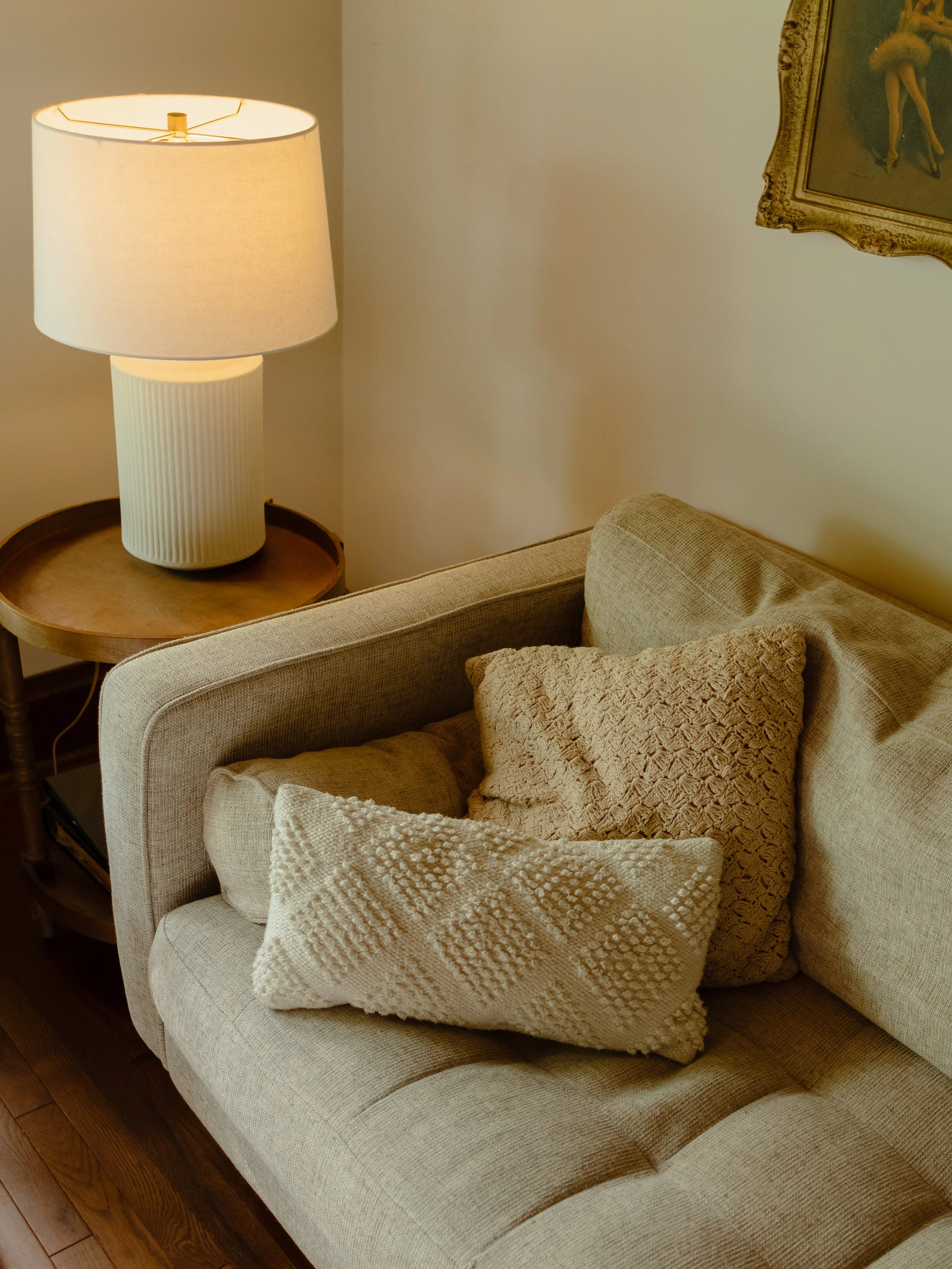 A beige upholstered sofa with two decorative pillows, placed next to a wooden side table with a lamp on top, in a cozy living room.