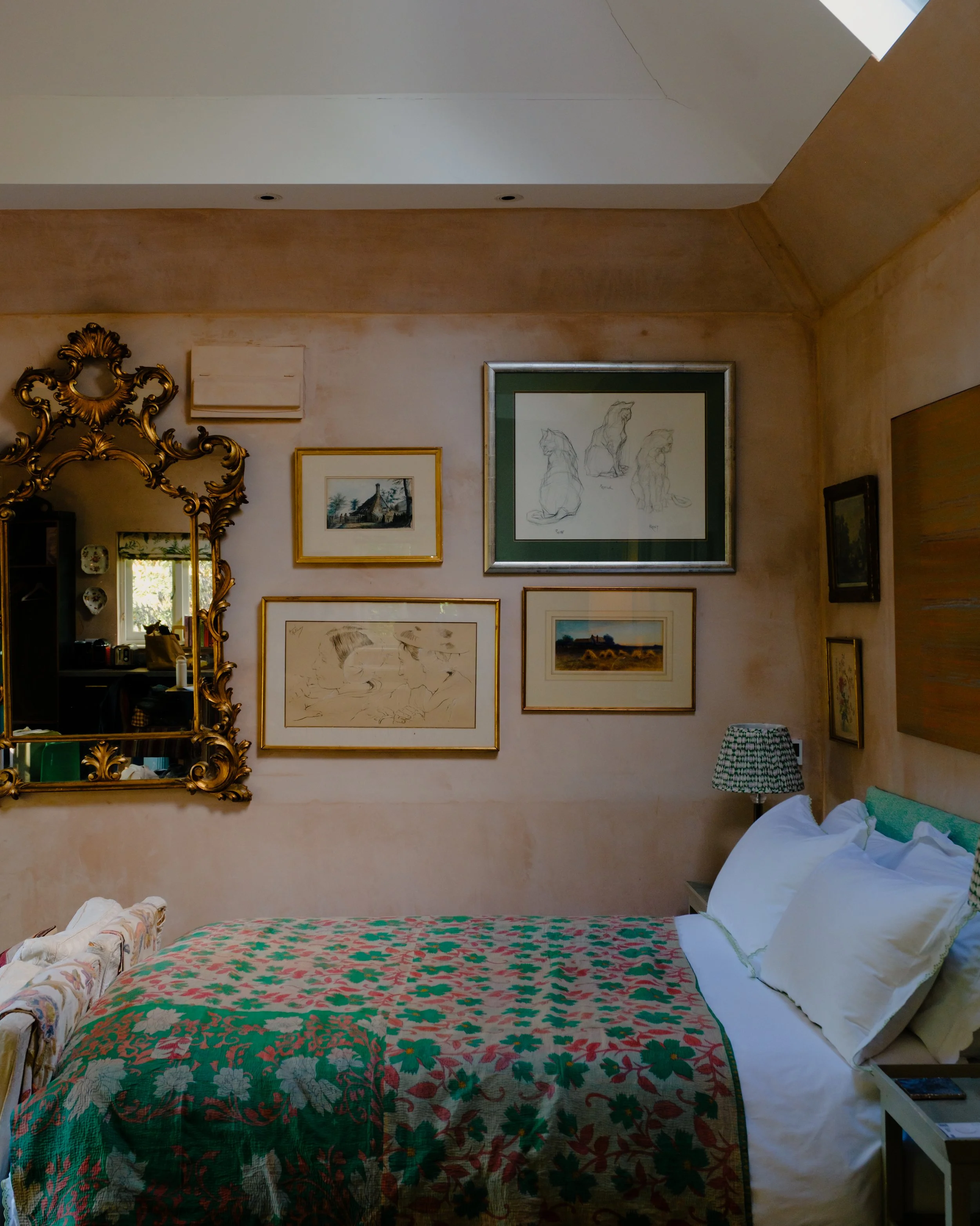 A bedroom with a bed covered in a green and red floral quilt, with white pillows and a bed skirt. The wall behind the bed features various framed artworks, including sketches of dogs, a landscape, and other pictures. There is a large ornate gold mirror on the left side, reflecting part of the room. A small table or nightstand partially visible on the right holds a patterned lampshade. The ceiling is beige with a crack, and there is a white wall with a crack near the ceiling.
