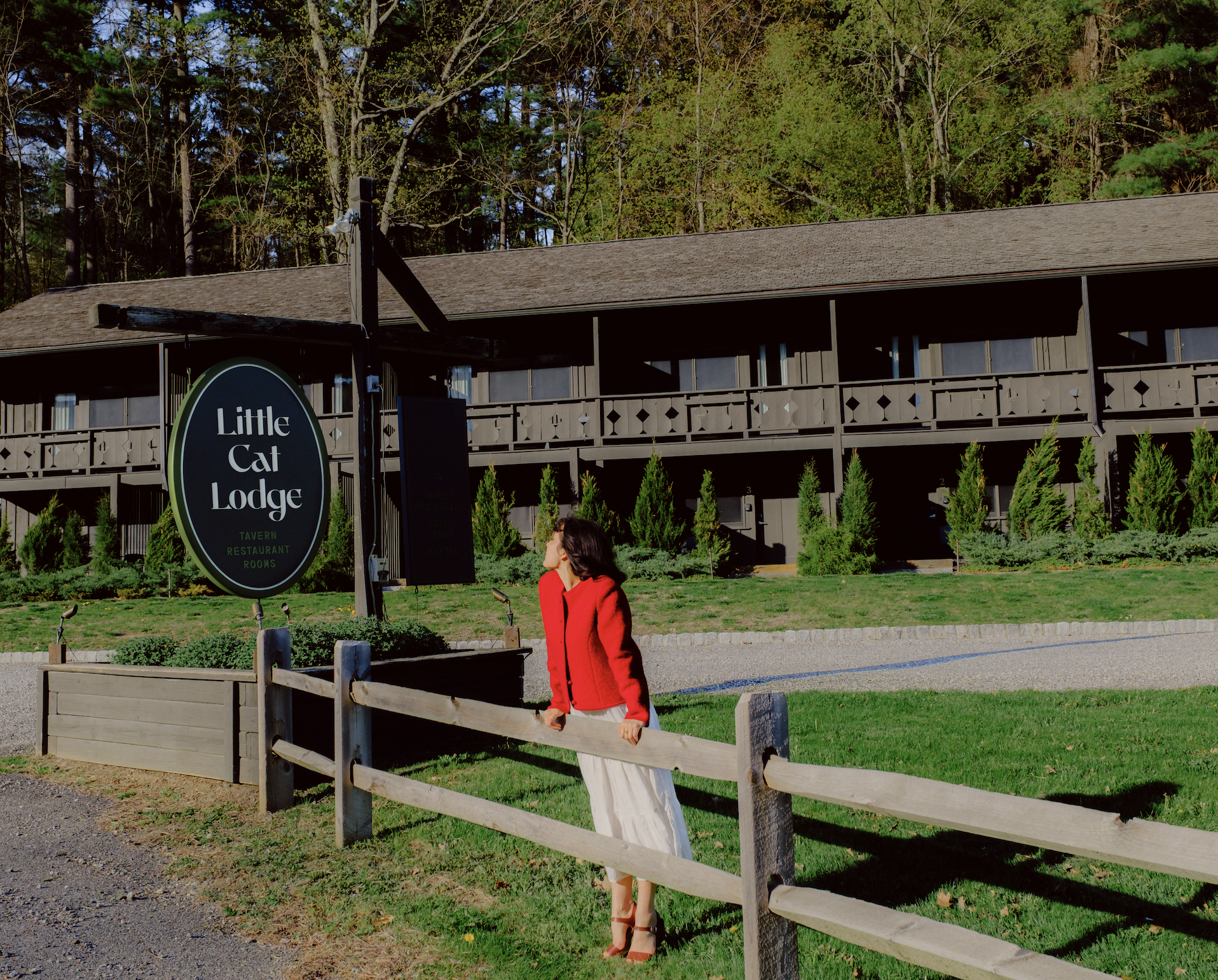 Woman in a red jacket standing outdoors near a wooden fence with a restaurant building and trees in the background.