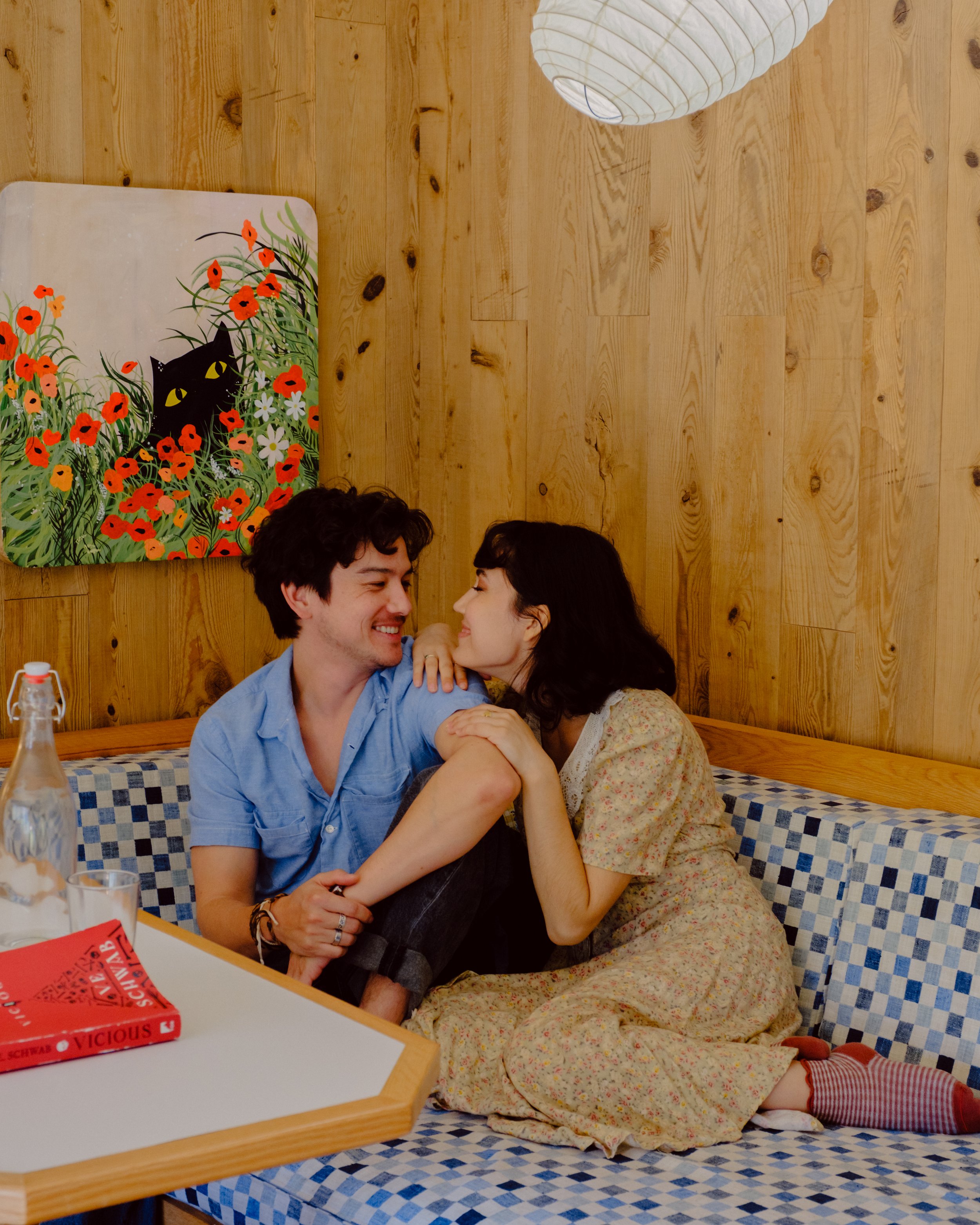 A man and woman sitting on a checkered booth in a restaurant, smiling and leaning close to each other, with a wooden wall and a decorative cat painting behind them.