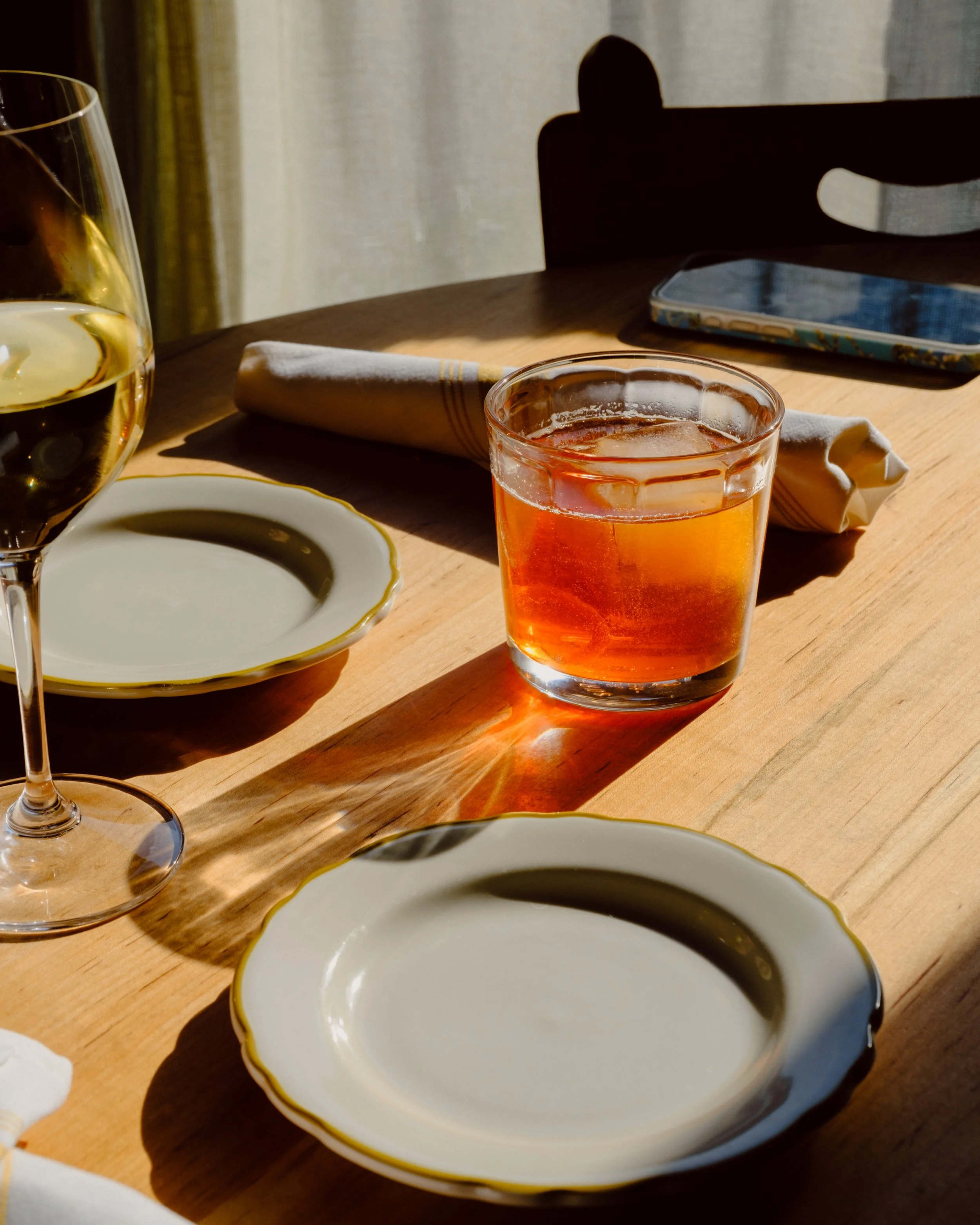 A dining table with a glass of white wine, an orange-colored drink in a glass, a white plate with a gold rim, a rolled napkin, and a smartphone on the table. Behind, there's a chair with a dark wooden backrest and a curtain.