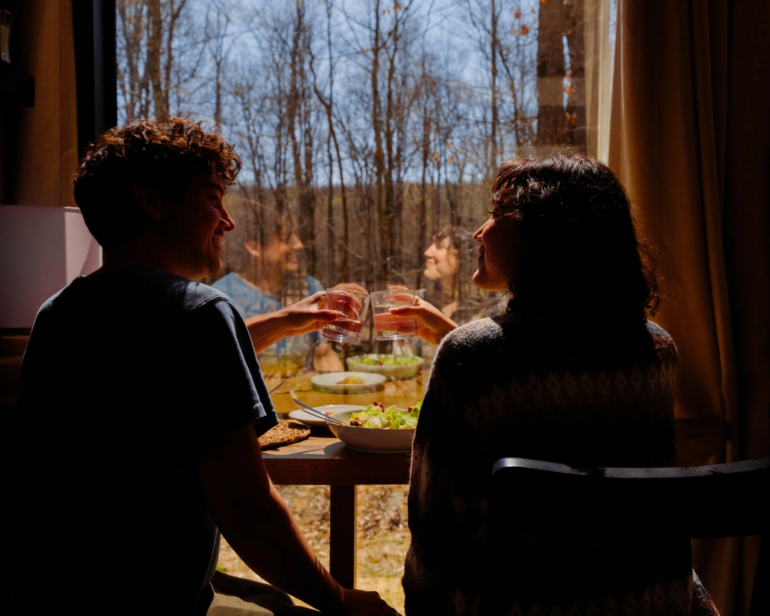Two people sitting indoors near a window, sharing drinks, with a background of trees outside