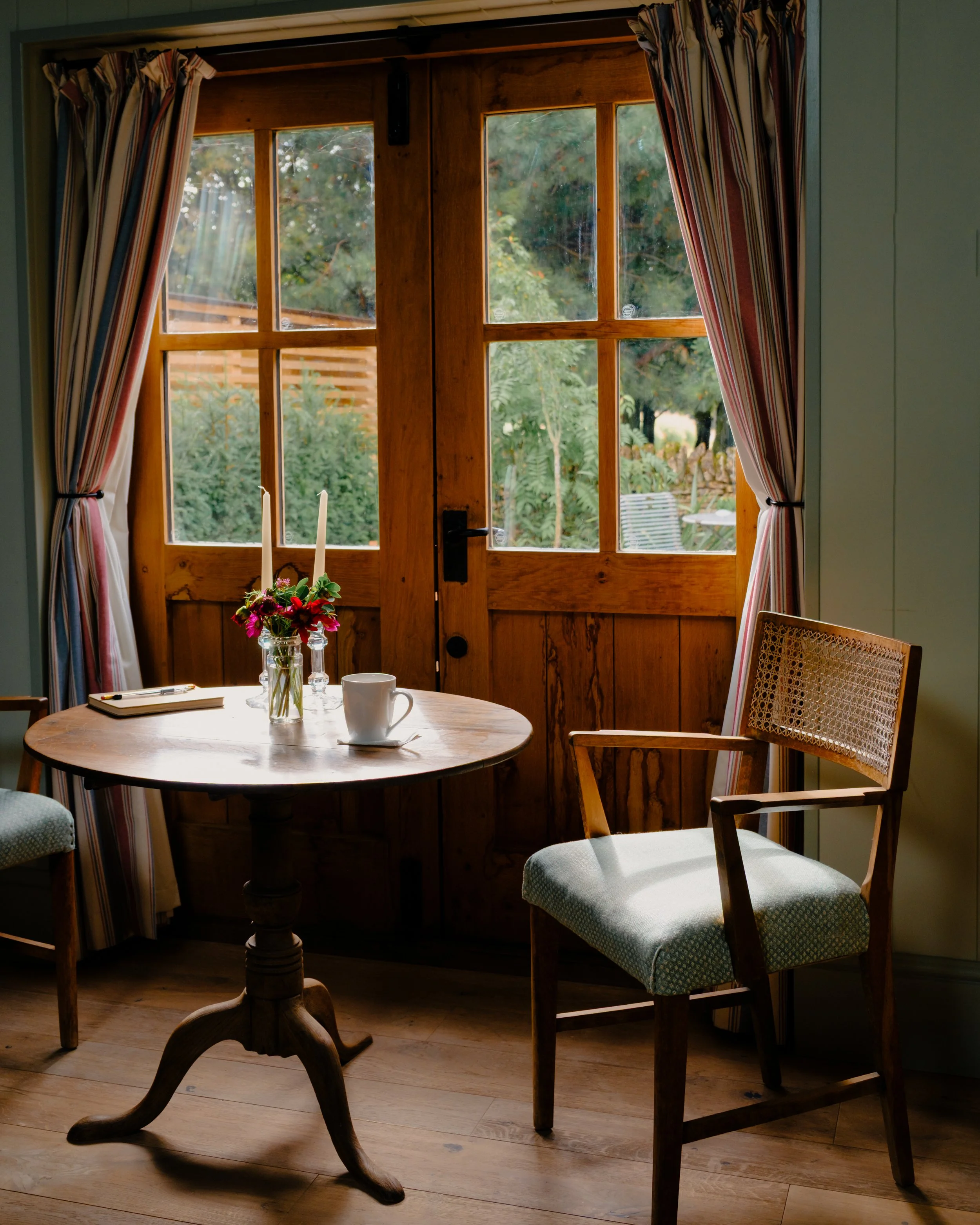 A cozy wooden interior room with a round table set for two, decorated with a small flower arrangement, candles, a notebook, and a cup. Large wooden-framed windows with striped curtains reveal a lush garden outside with trees and seating.