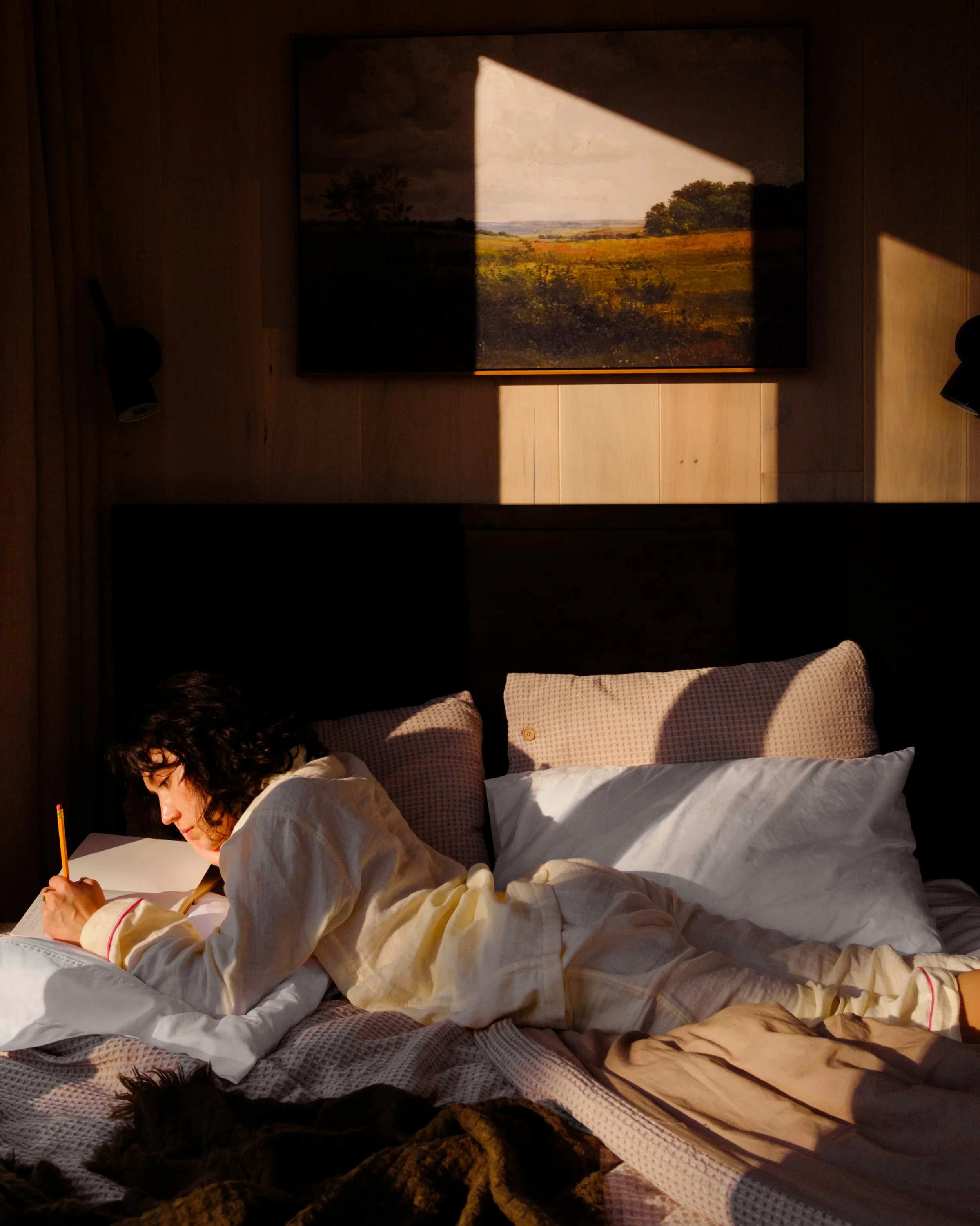 A woman lying on her side in bed, writing in a notebook, with sunlight casting shadows through a window.