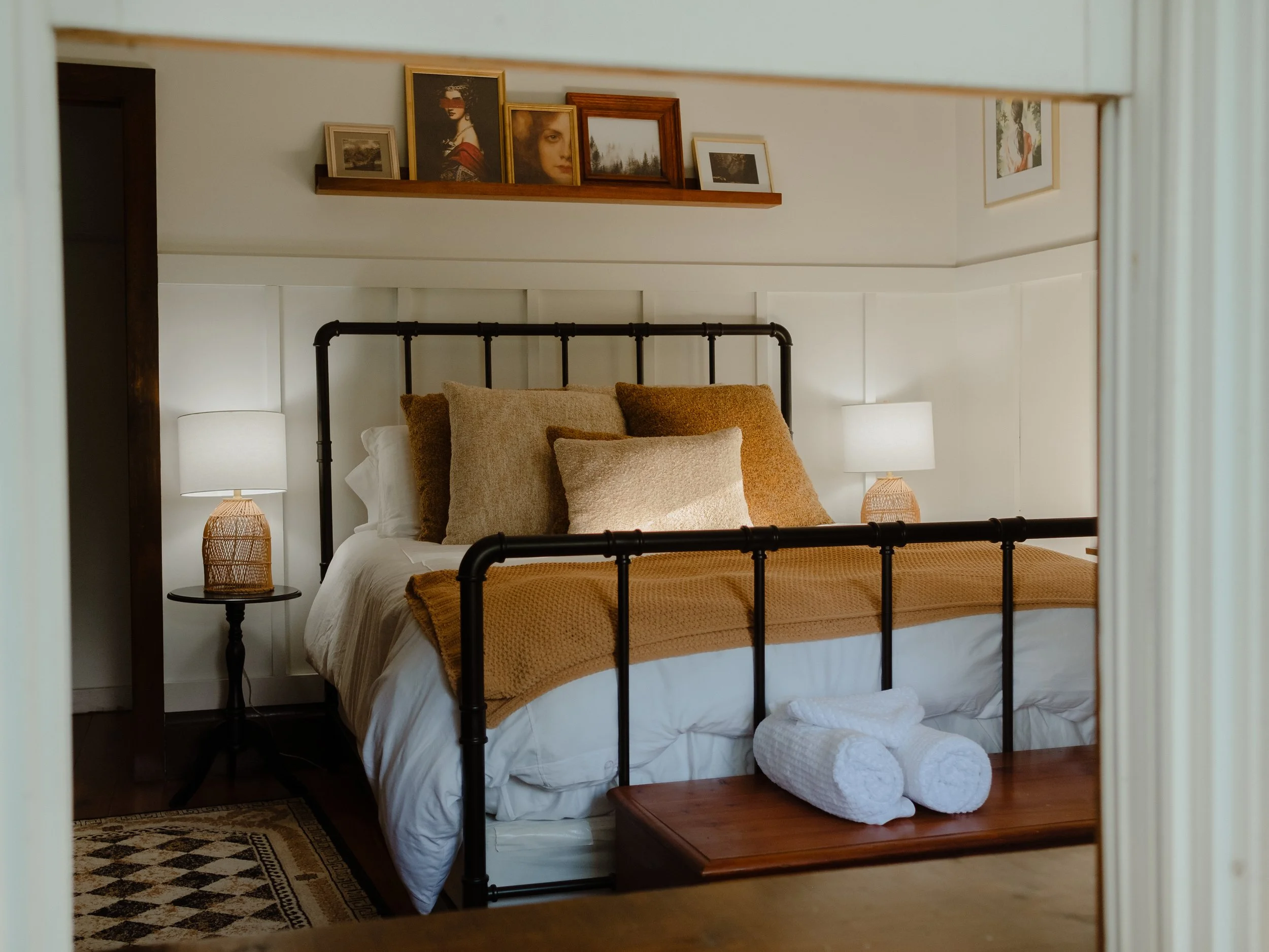 A cozy bedroom with a black metal bed frame, tan and beige pillows, a white comforter, and a wooden bench at the foot. Two nightstands with woven lamp shades flanking the bed, topped with beds, and a shelf above displaying framed art and photographs,