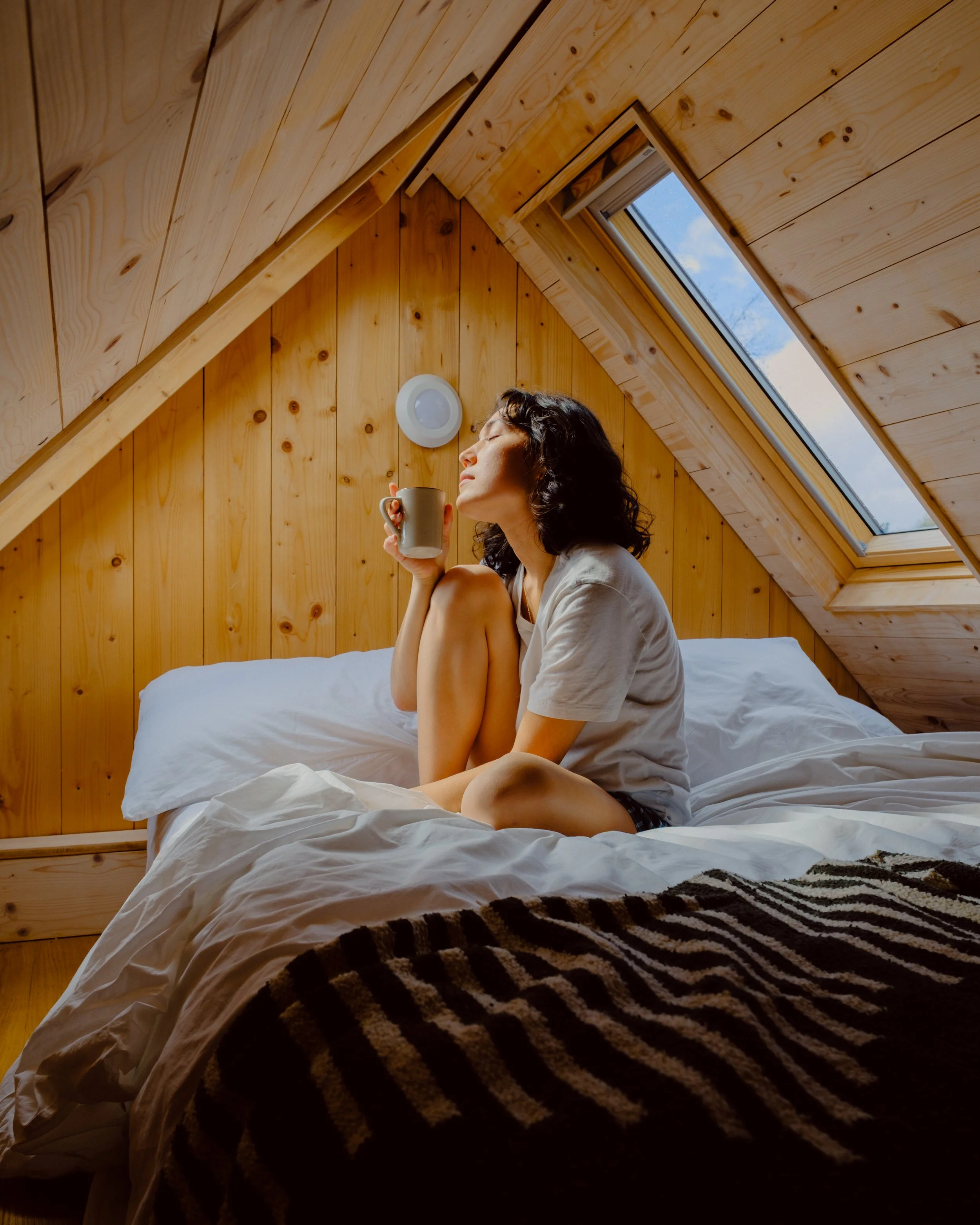 A woman with dark curly hair is sitting on a bed in a wooden room with a sloped ceiling and a skylight window. She is holding a mug and appears to be enjoying the sunlight coming through the window.