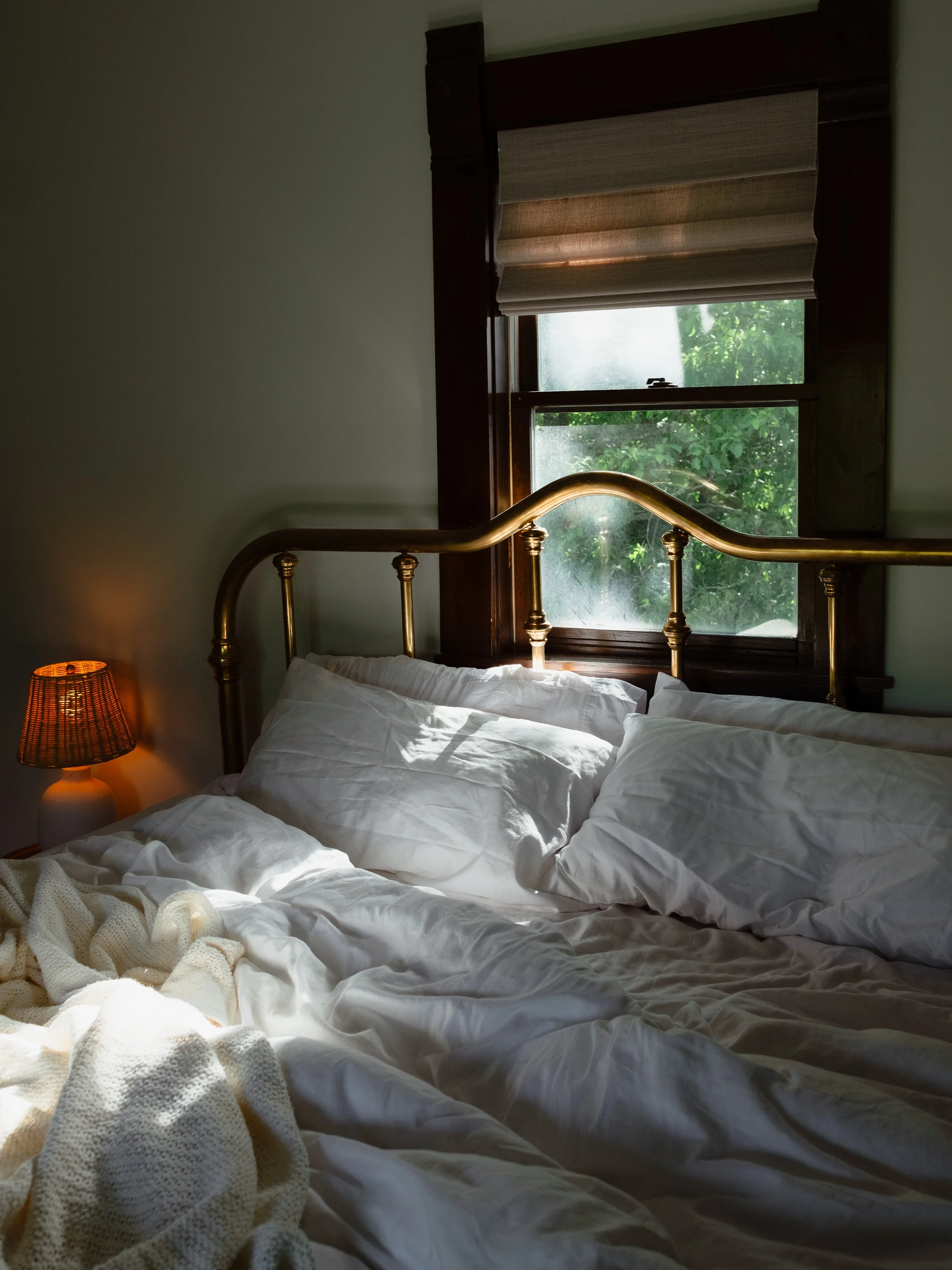A bedroom with a brass bed, white bedding, a window with a wooden frame and blinds, and a small lamp emitting warm light.