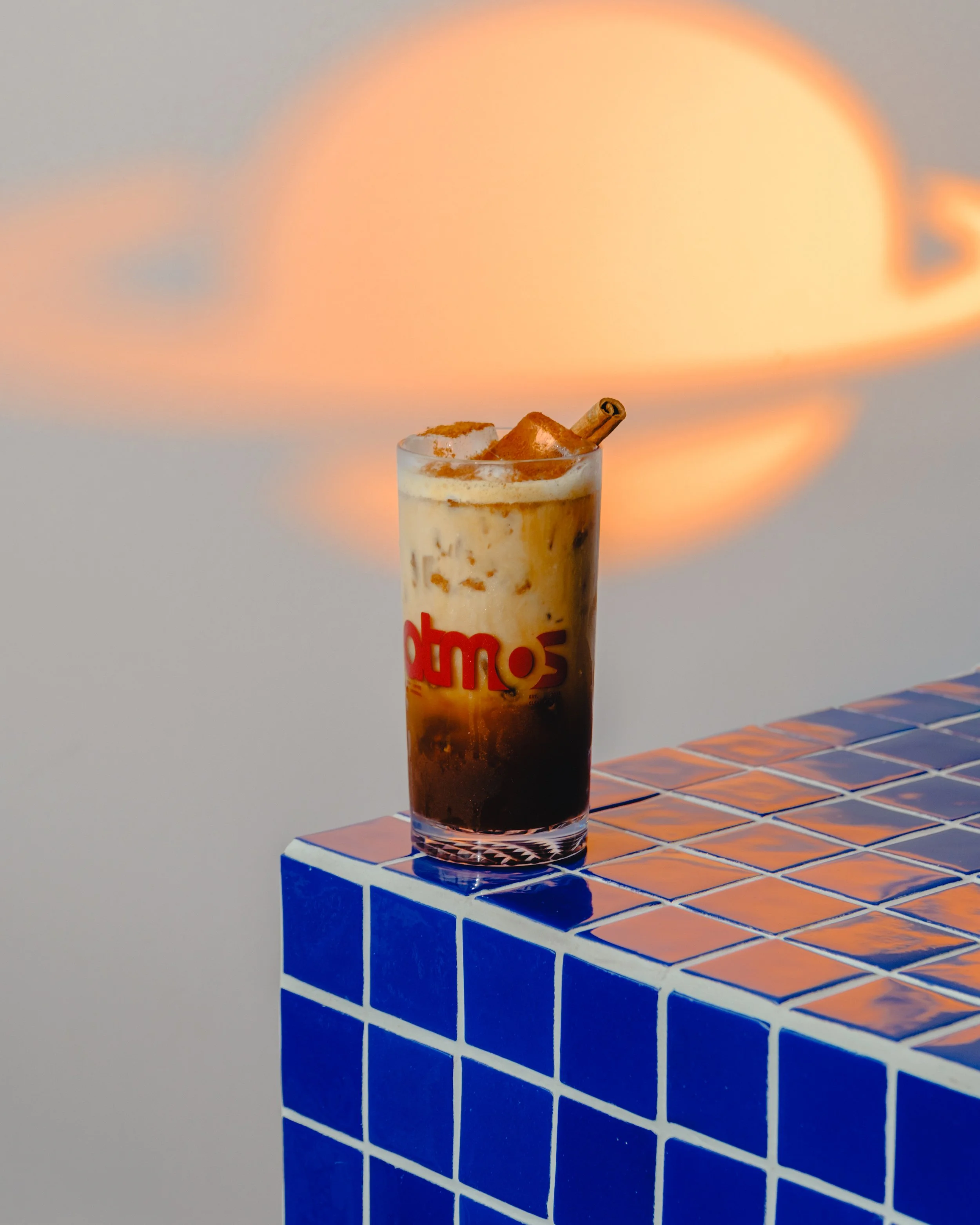 A tall glass of iced coffee with cinnamon and a cinnamon stick, placed on blue tiled table with orange and blue pattern, with blurred sunset sky background.