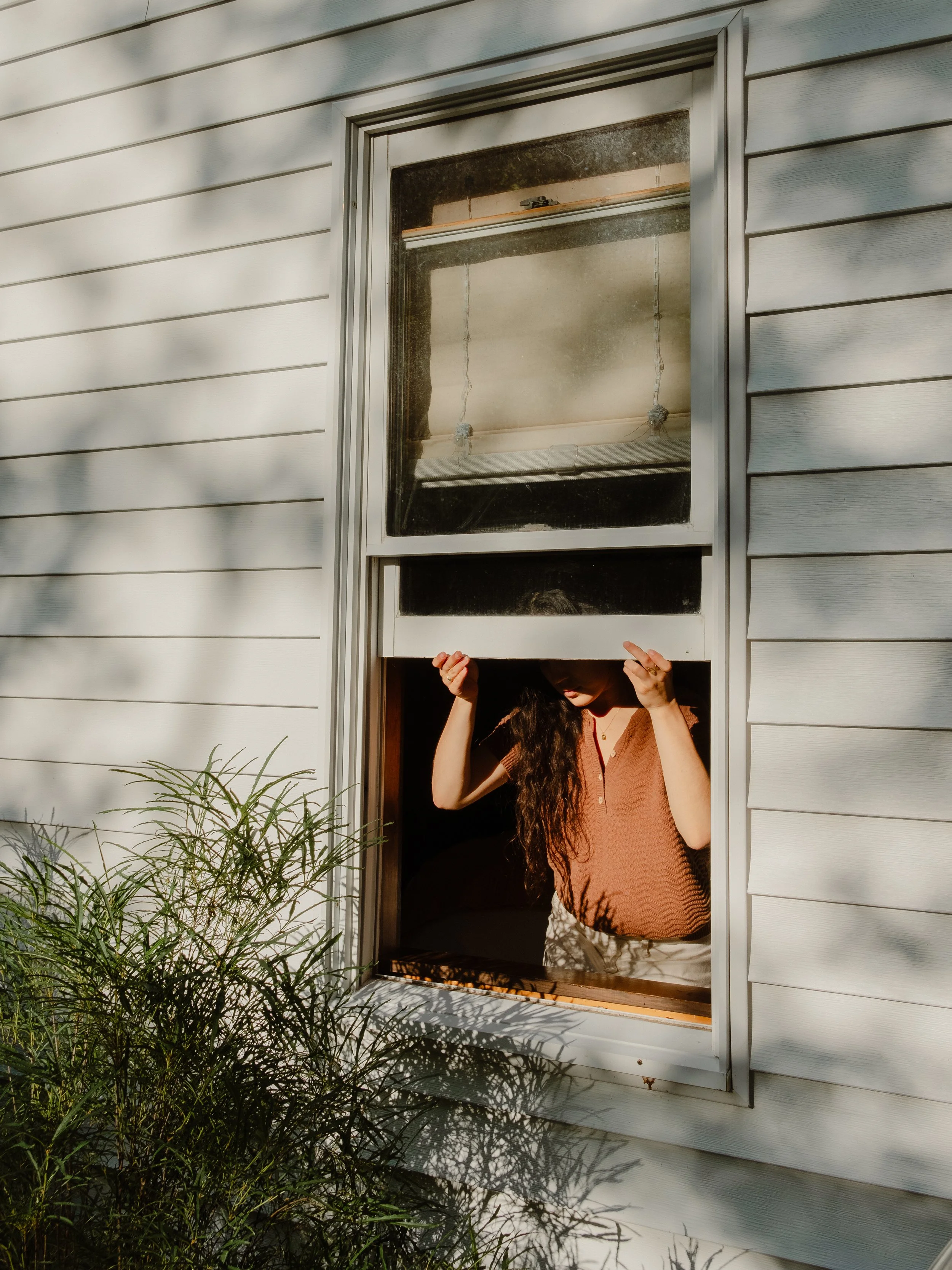 A person with long dark curly hair is looking out of a bathroom window, holding the top of the window frame. There are white horizontal siding on the exterior wall and some green plants in the foreground.