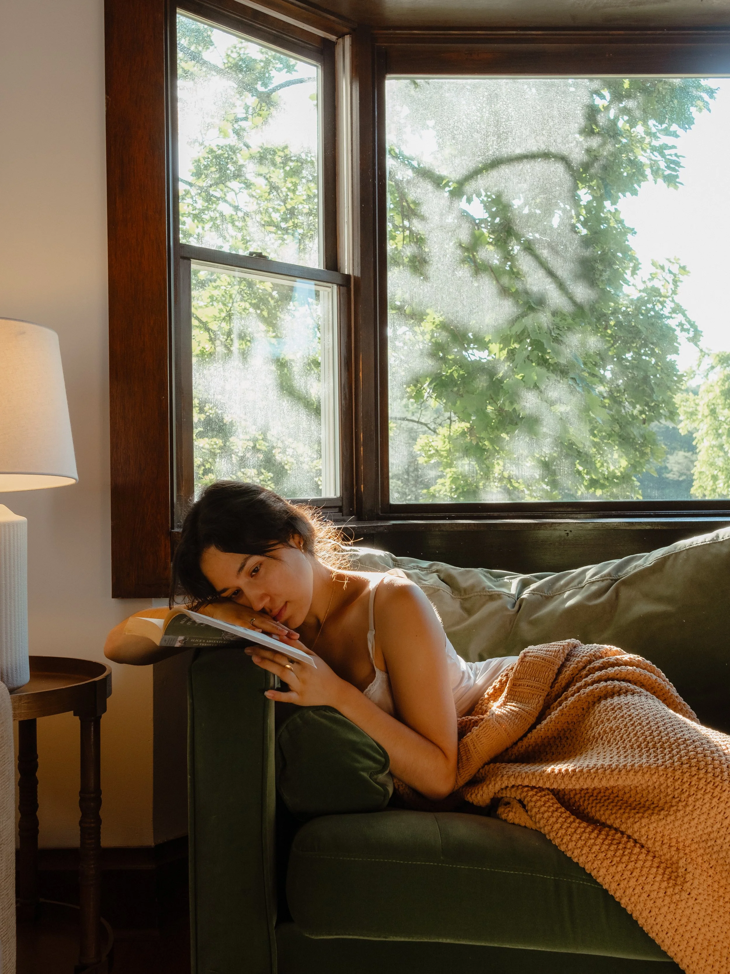 A woman with dark hair is lying on a green sofa, reading a book near a window with trees outside. She is covered with a knitted orange blanket and is resting her head on a pillow, with sunlight coming through the window.
