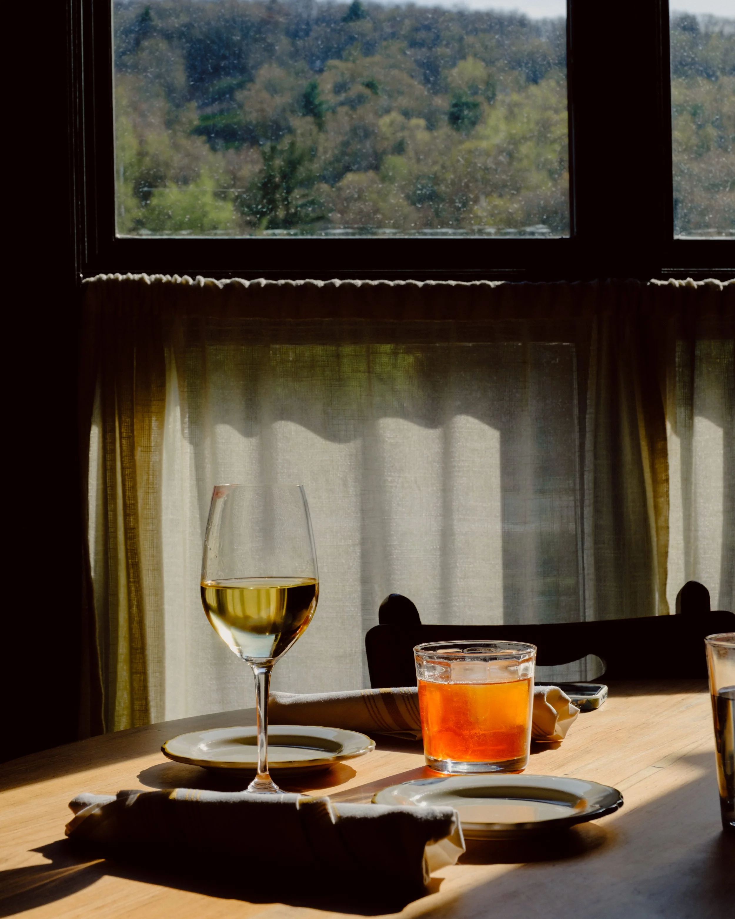 A glass of white wine, a glass of orange juice, and a glass of water on a wooden table near a window with curtains, sunlight casting shadows.