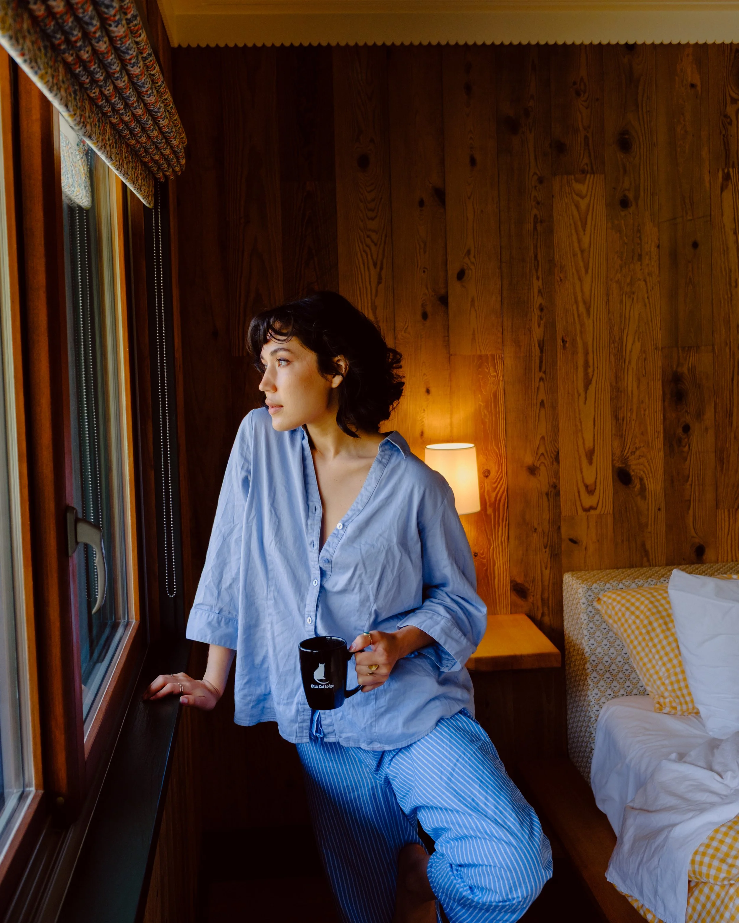 A woman with dark, short, curly hair looking out a window while holding a black mug in a cozy wooden-paneled room. She wears a light blue button-up shirt and striped pants.