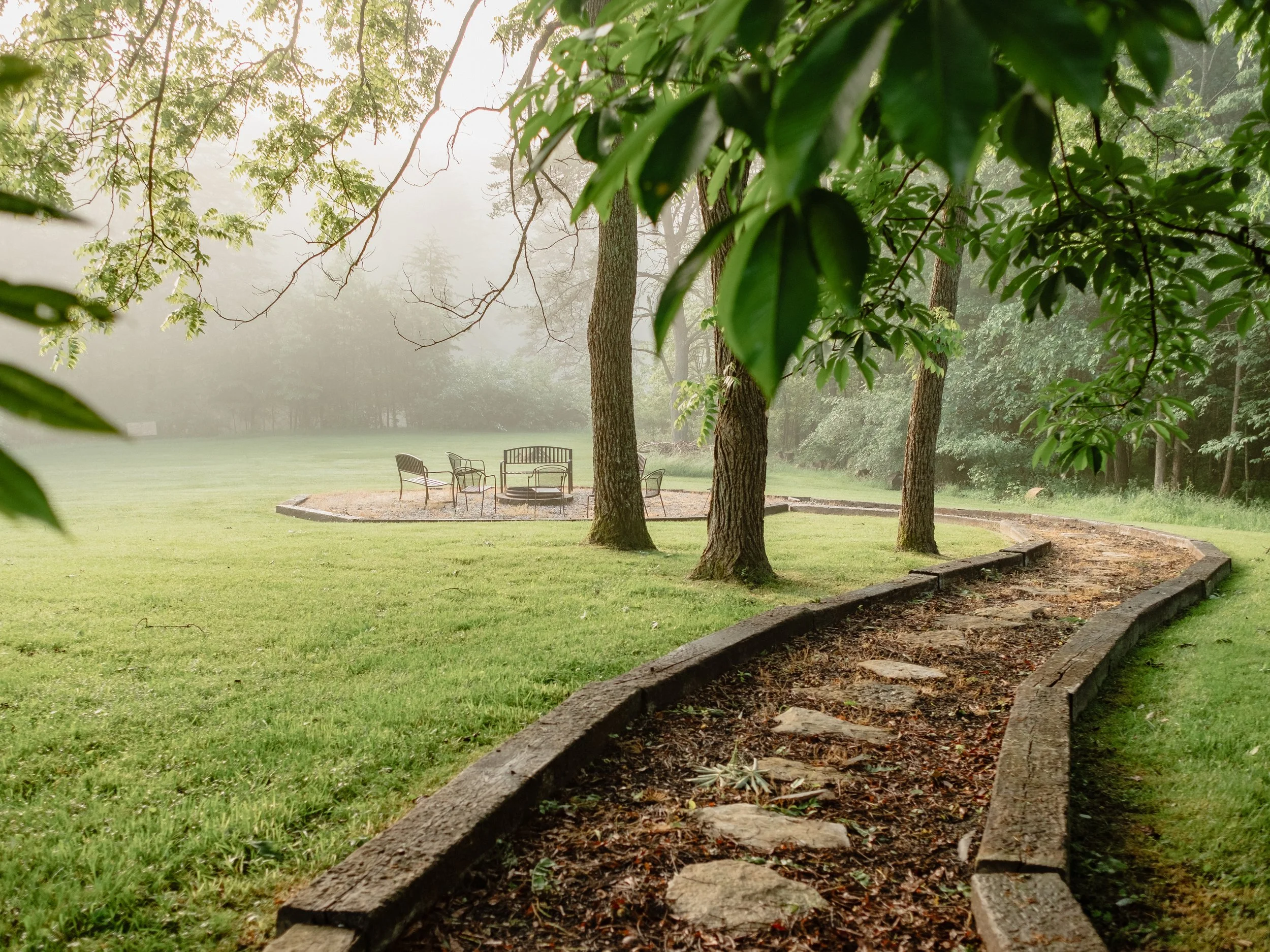 A park scene with surrounded by trees, a fire pit with chairs around it, and a grassy area with a gravel walkway and a curved stone border.
