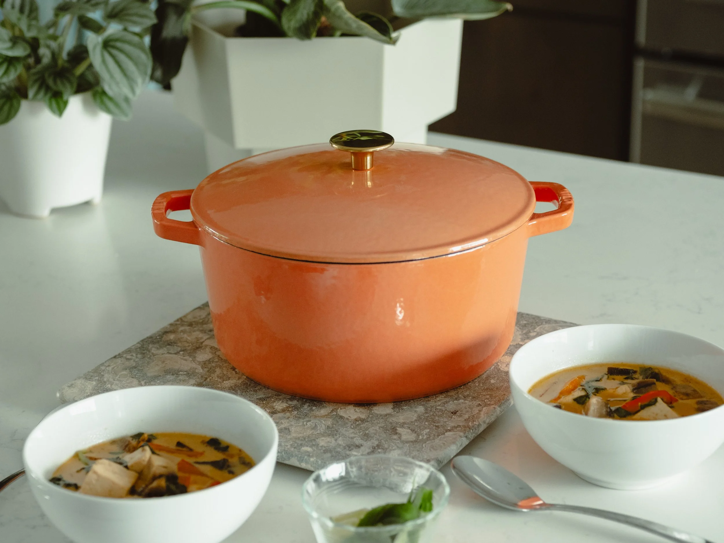 Orange ceramic pot with a lid on a kitchen countertop, surrounded by bowls of soup and a potted plant in the background.