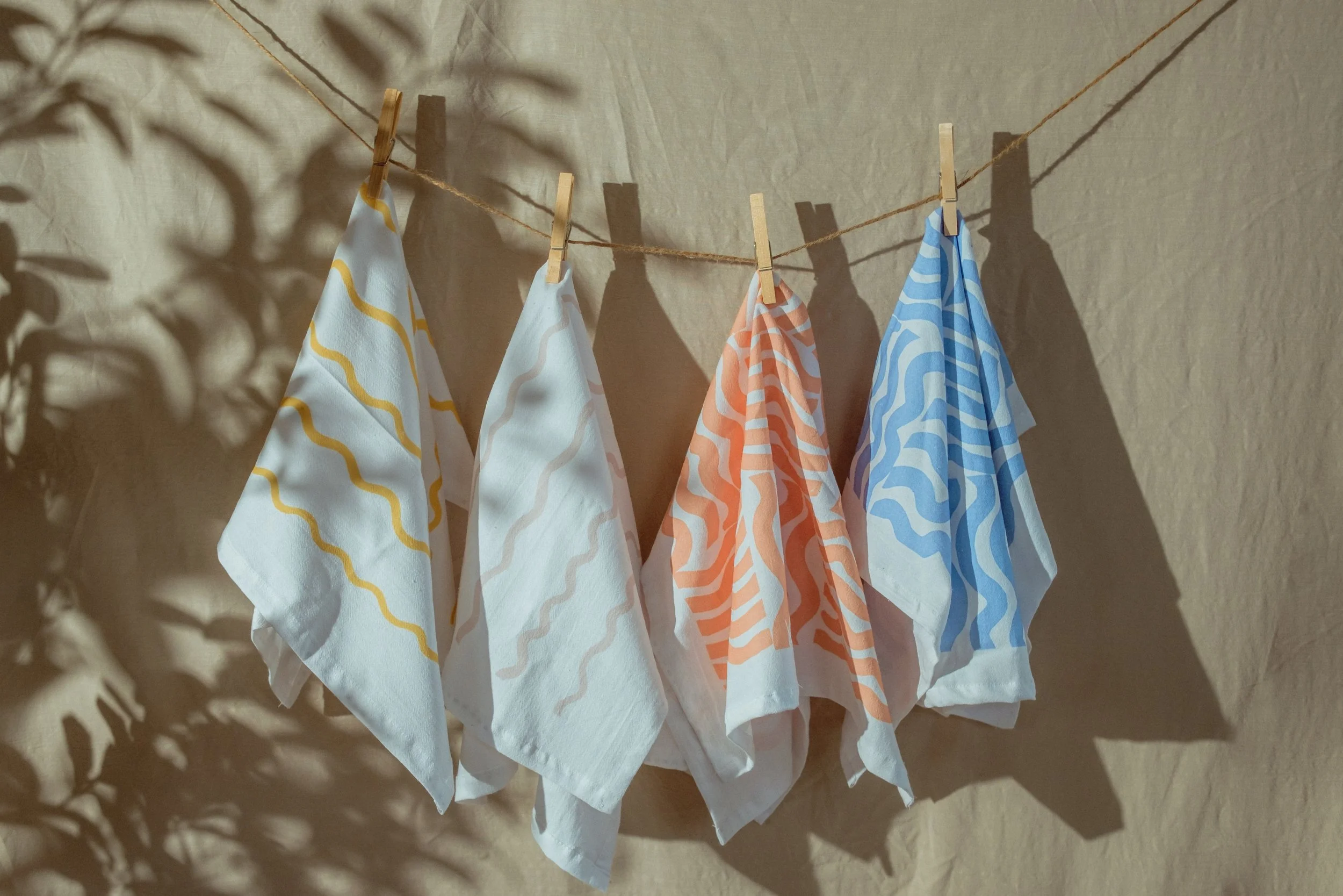 Colorful patterned cloths hanging on a clothesline secured with small wooden clothespins, casting soft shadows on a plain wall.