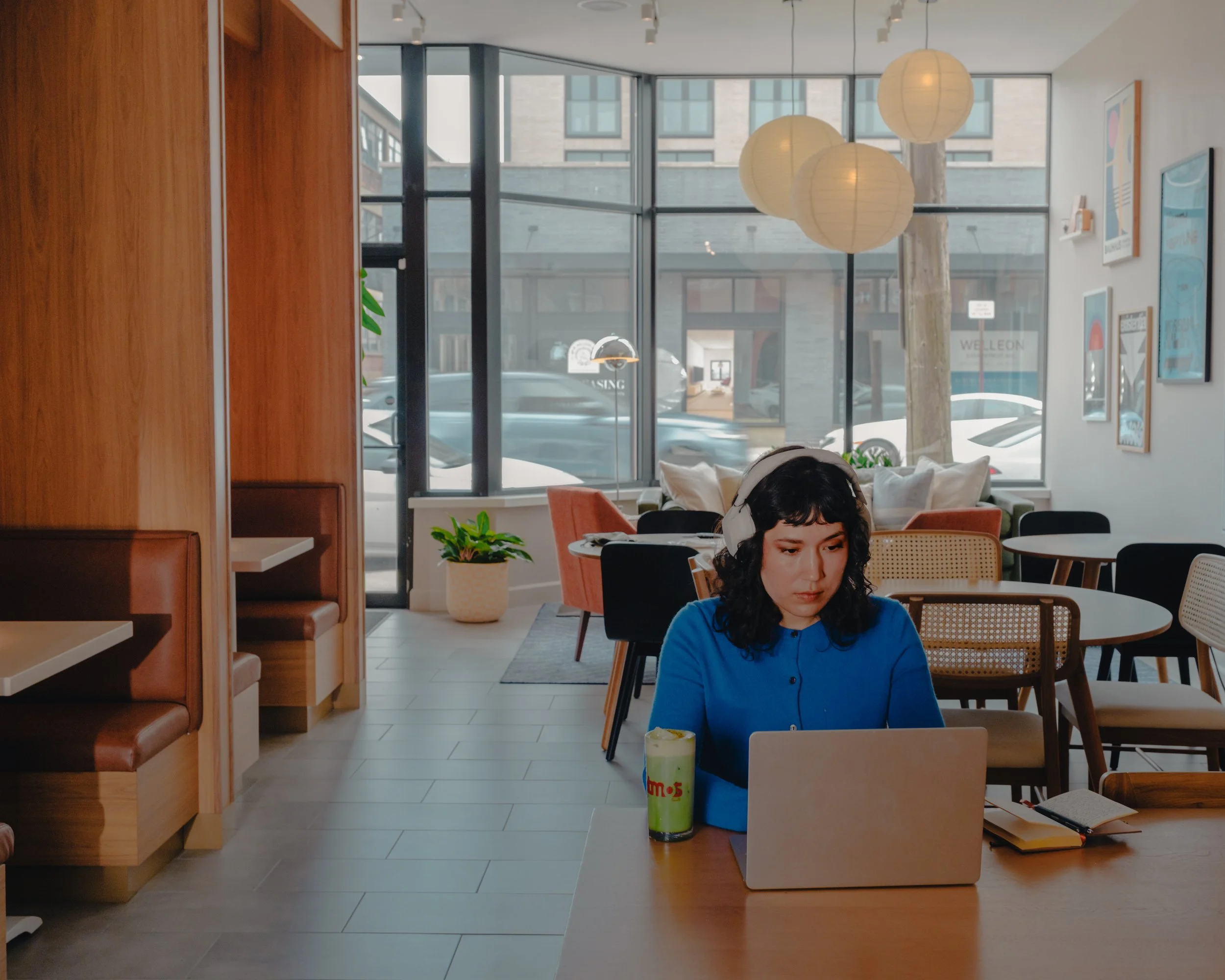 Interior of a modern cafe or restaurant with large windows, hanging round paper lamps, and framed artwork on the walls. A woman with dark, curly hair and a blue shirt is seated at a table, looking down, with colorful drinks and notebooks nearby.