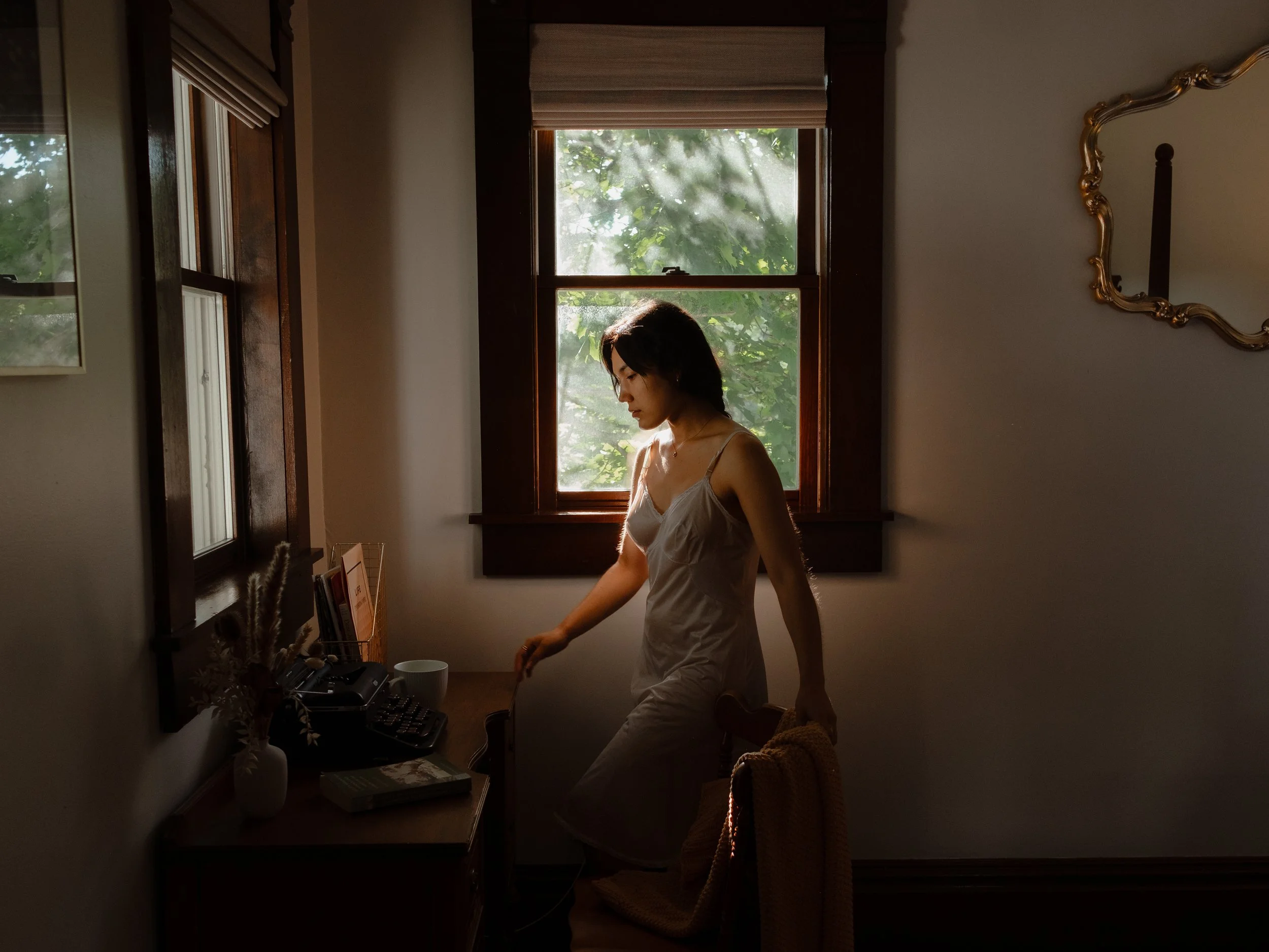 A woman in a white dress standing near a desk with office items, backlit by a large window with green foliage outside, in a cozy room