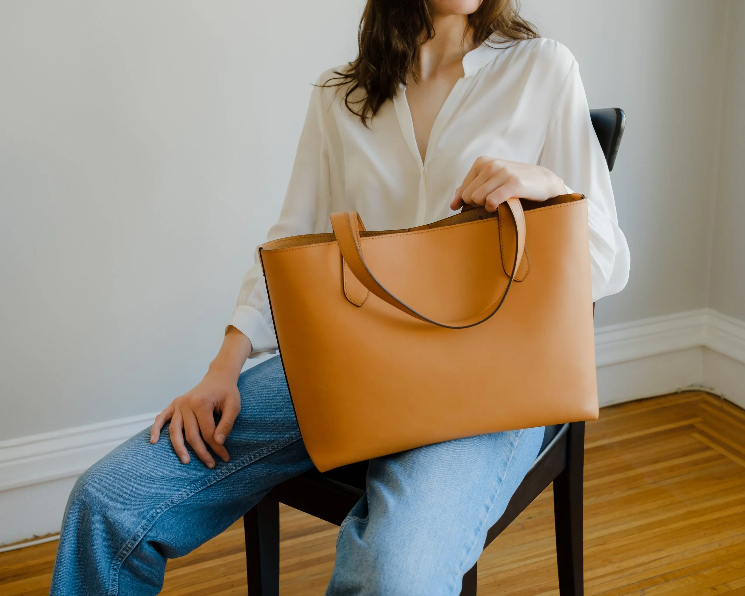 A woman sitting on a black chair in a room with wooden flooring, wearing a white blouse and blue jeans, holding a large tan handbag on her lap.
