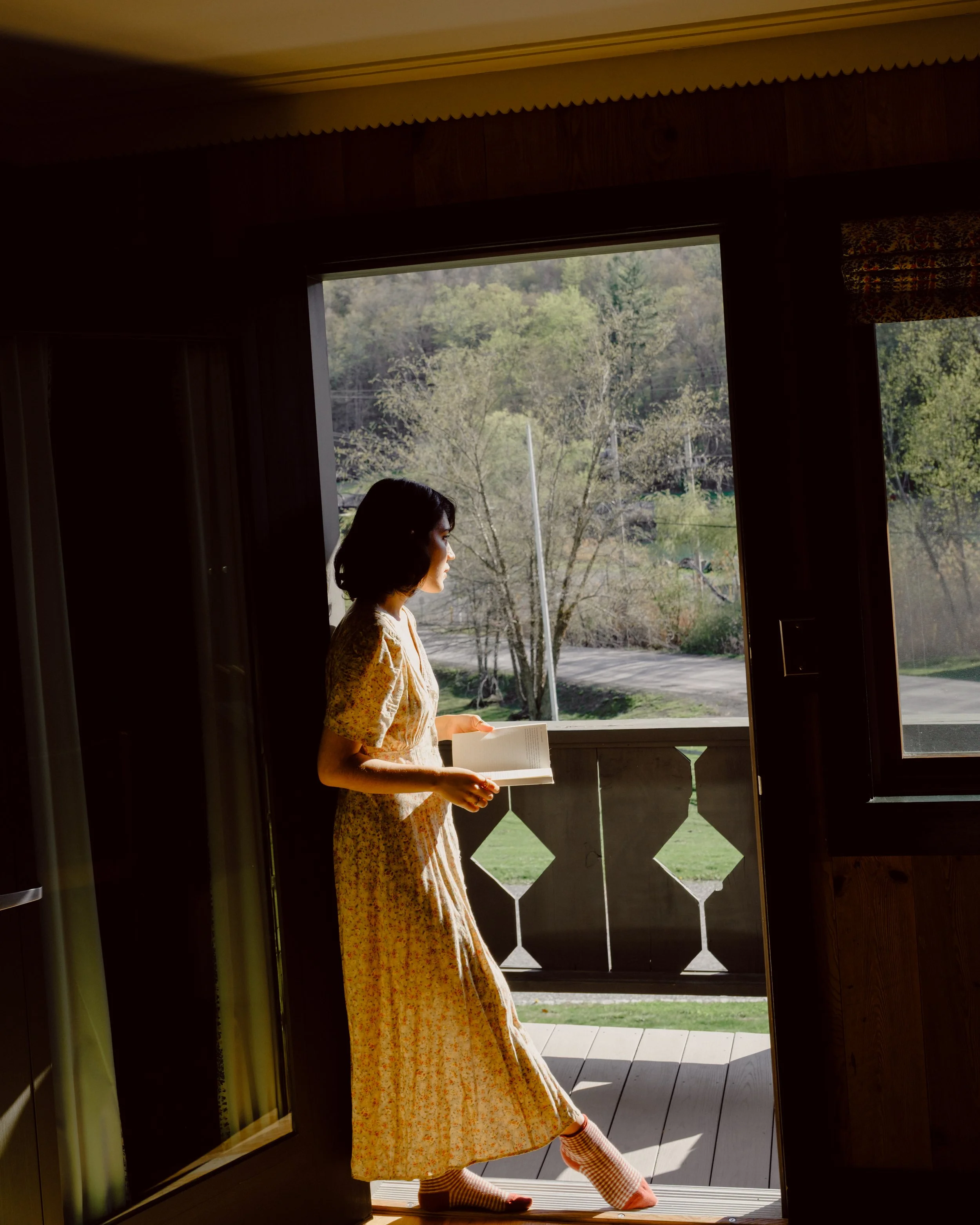 A young woman with dark hair stands at a doorway, holding a book, with the outdoors visible through the open door, revealing trees and a sidewalk in a rural or suburban setting.