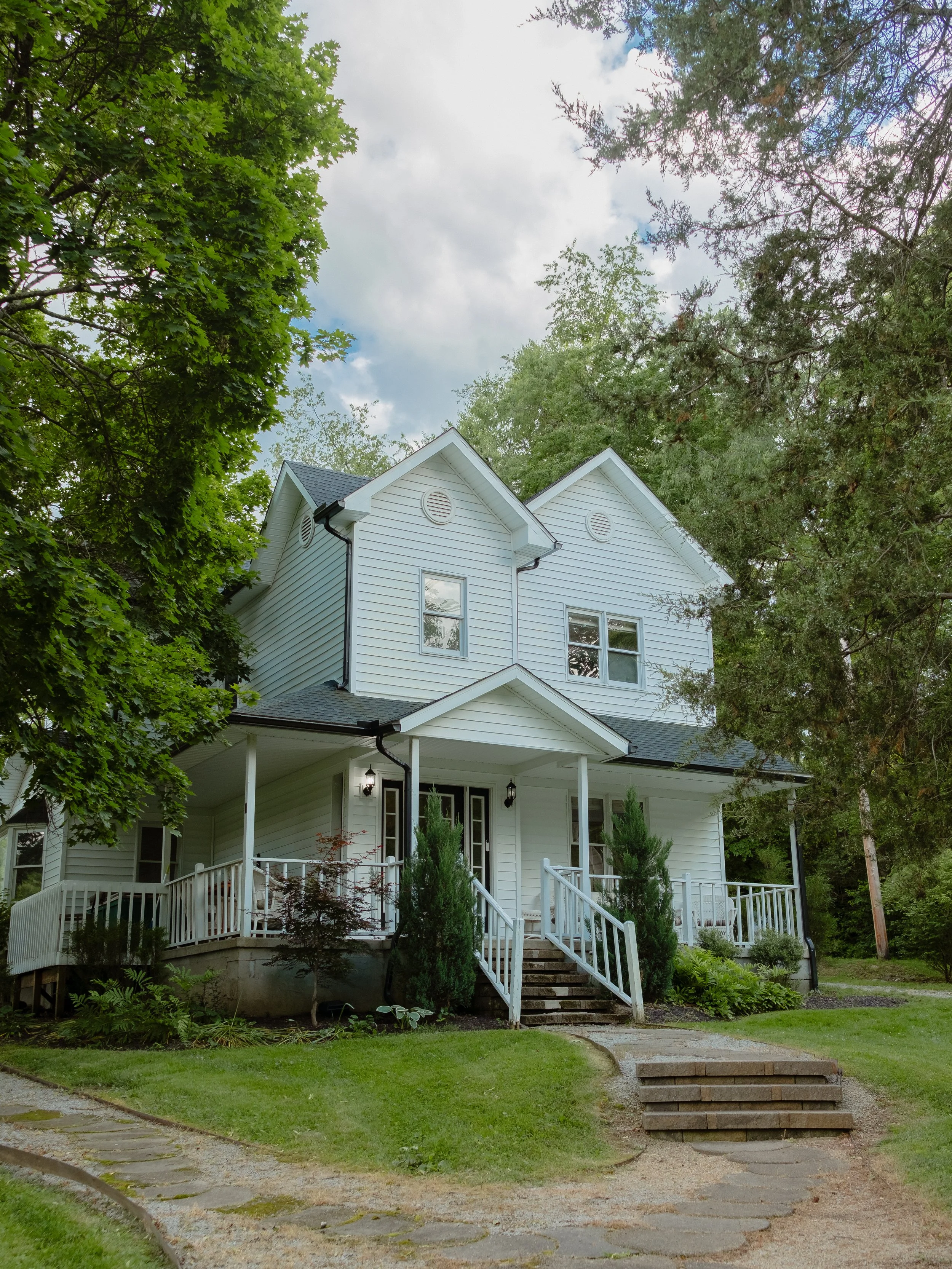 A white two-story house with a front porch surrounded by greenery and trees, with steps leading up to the entrance and a gravel pathway in front.