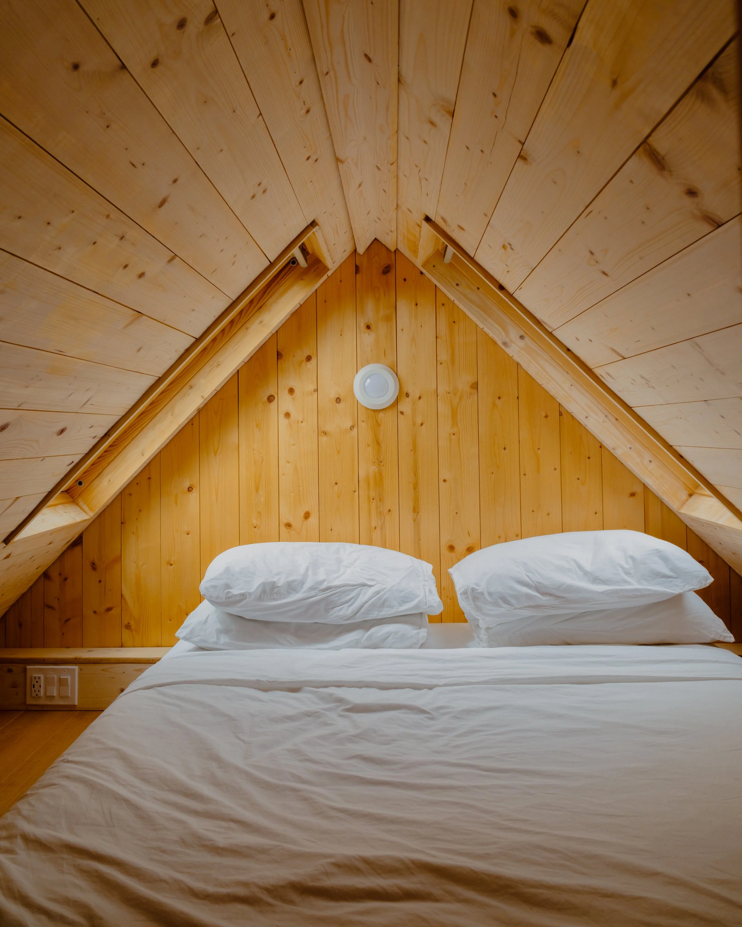 A bed with white sheets and piles of pillows against a wooden-paneled wall in a cozy attic bedroom.