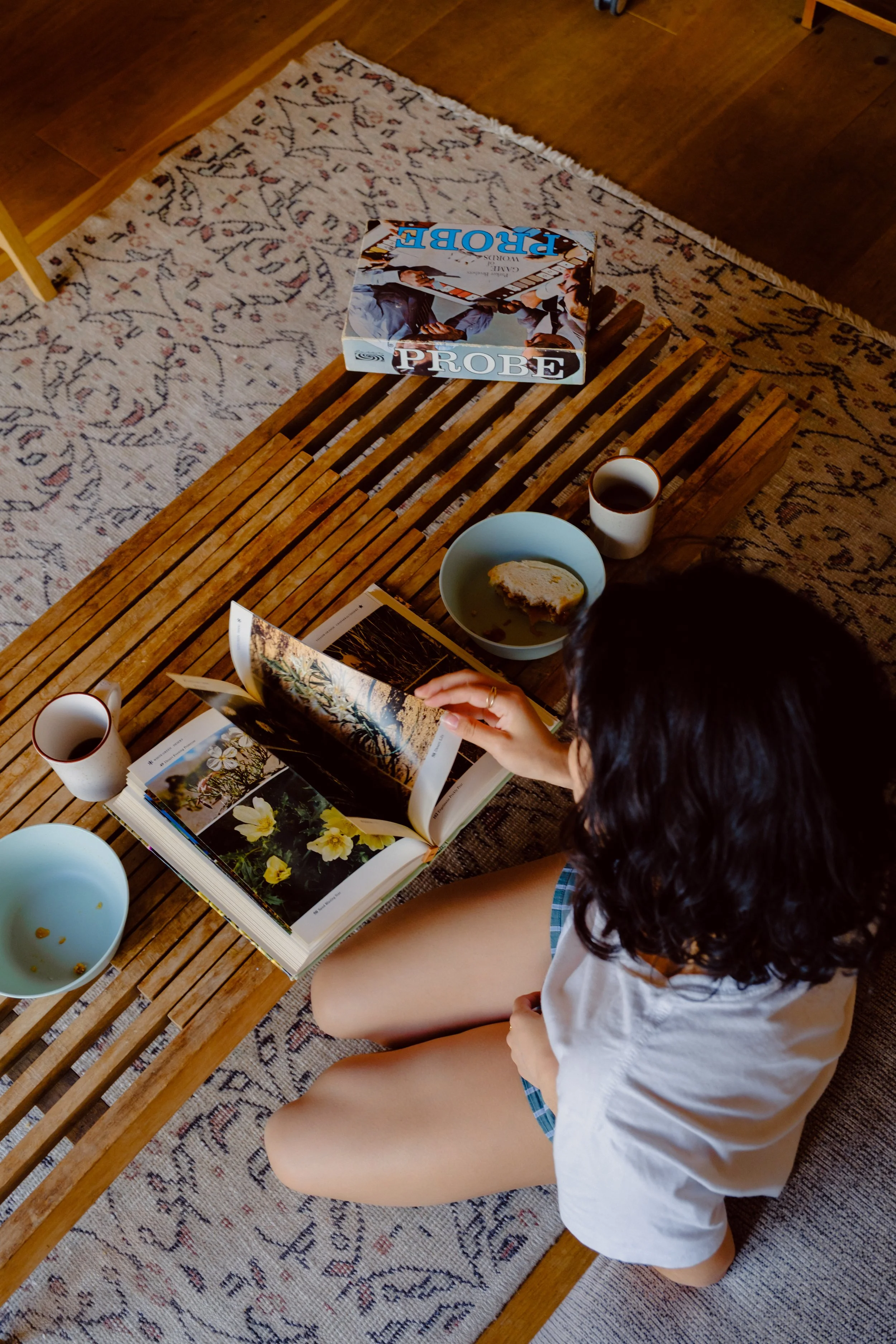 Person with long dark hair reading a large book with flower photographs, sitting on a wooden bench with a patterned rug underneath, with a cup of coffee, a partially eaten piece of bread, and a magazine on the table.
