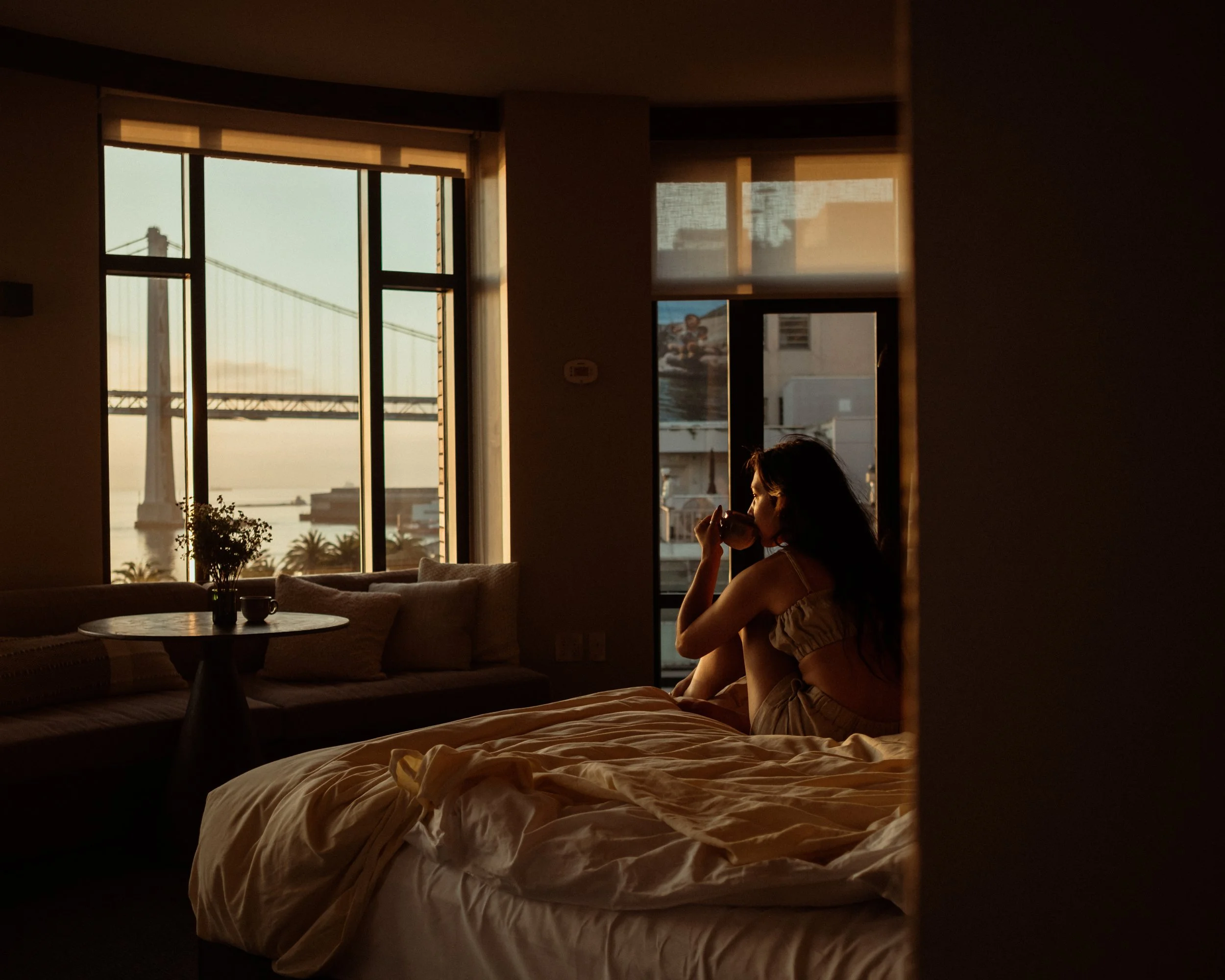 A woman sitting on a bed drinking from a cup, with large windows overlooking city buildings and the bridge at sunset.