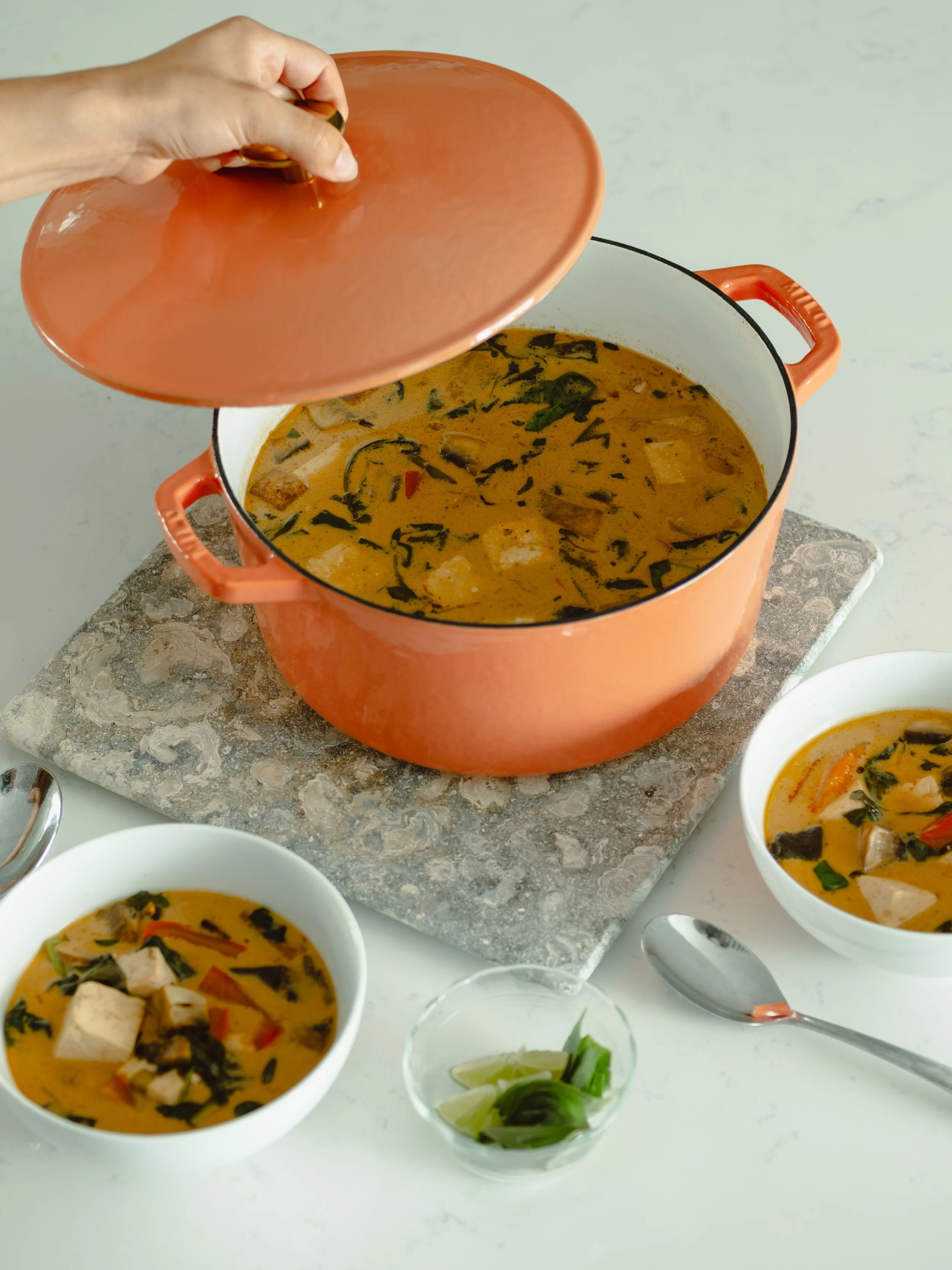 A pot of Asian soup with tofu, vegetables, and broth, with three white bowls of the soup and a small bowl of lime wedges and herbs nearby.