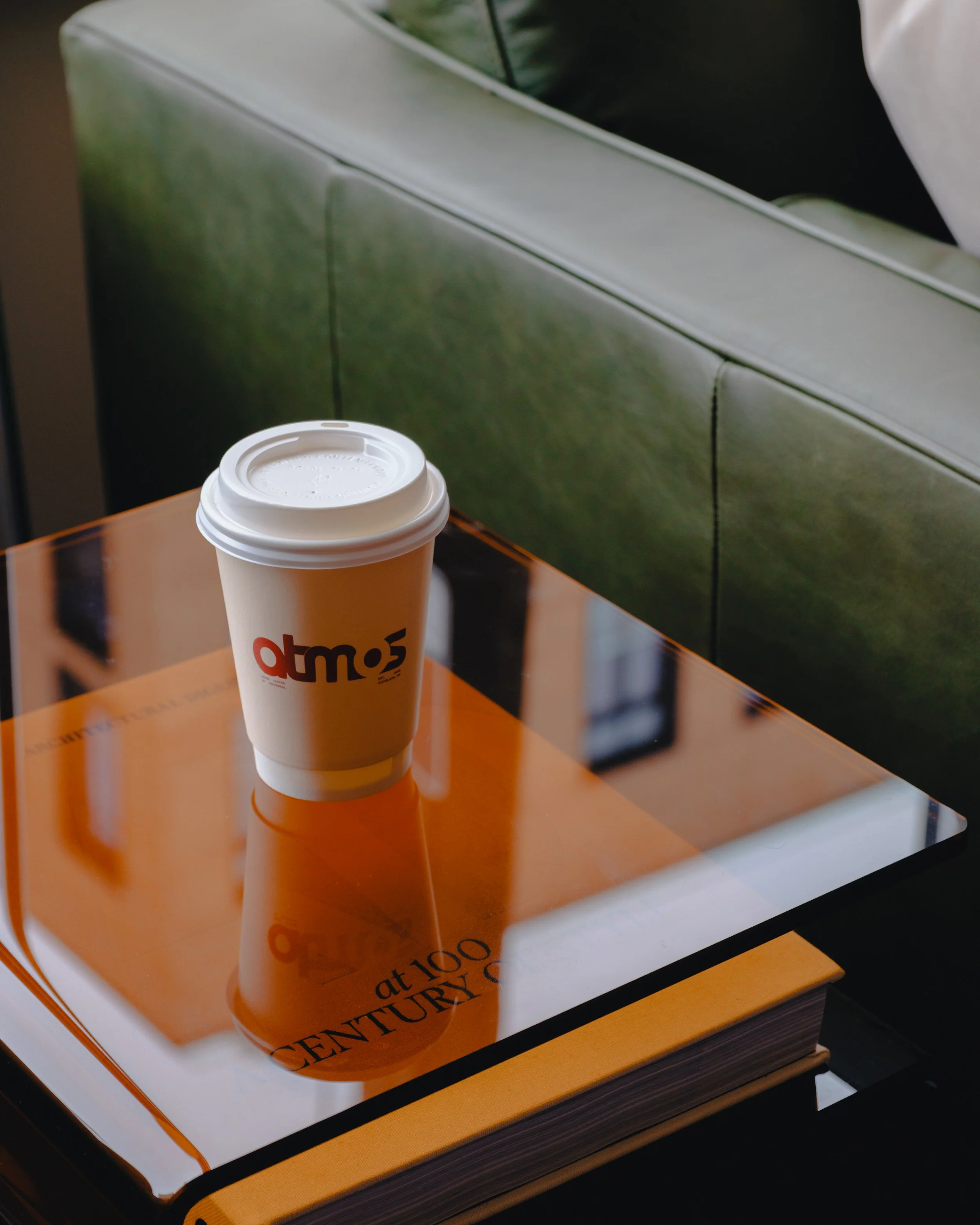 A disposable coffee cup with a white plastic lid placed on a glossy orange and white table, with a green seat or wall in the background.