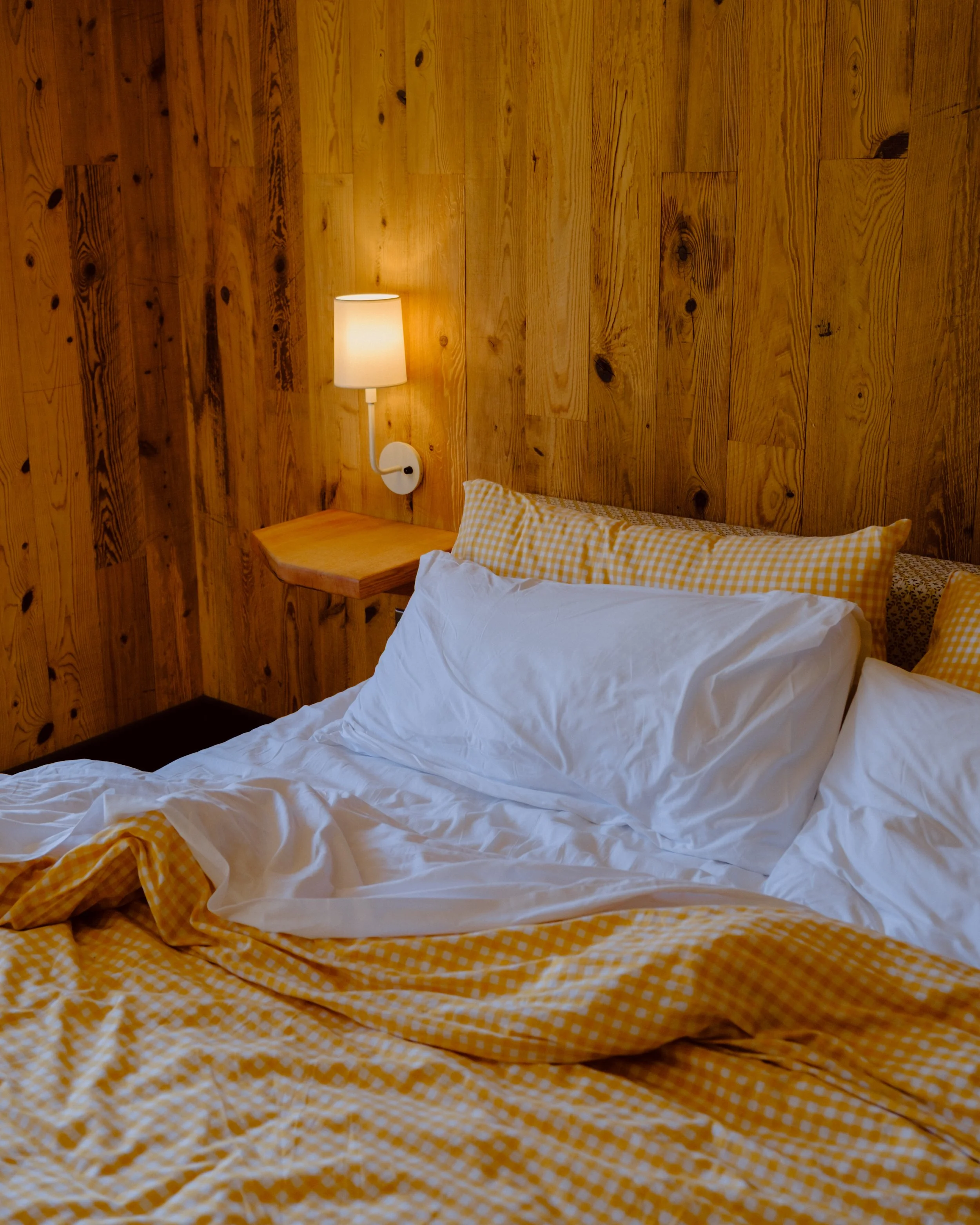 A cozy bedroom with a wall covered in wooden panels. There is a white wall-mounted lamp turned on next to a bed with a white duvet and yellow checkered pillows and bedspread. A small wooden bedside shelf is attached to the wall beneath the lamp.