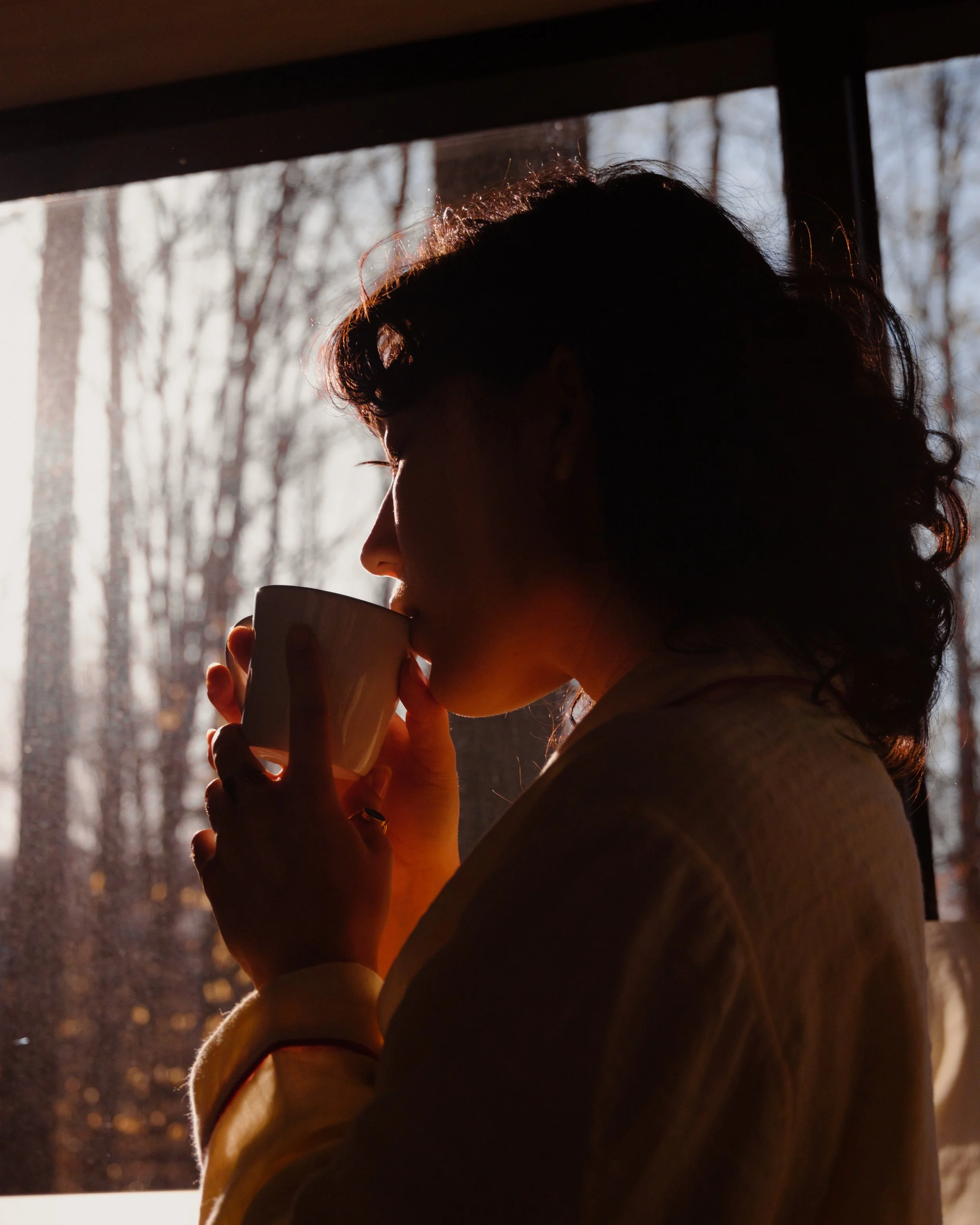 Person with curly hair drinking from a mug indoors, with a background of blurred trees visible through a window, backlit by sunlight.