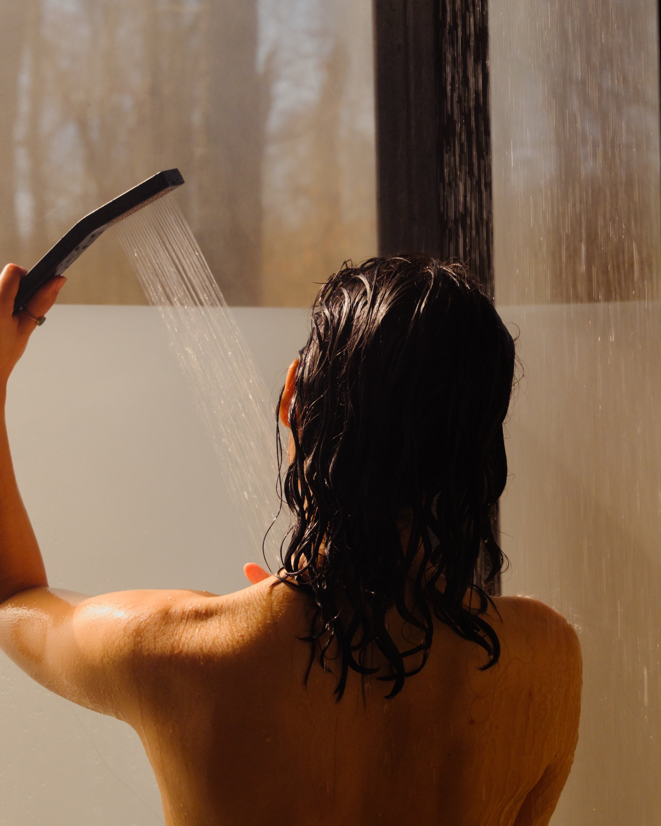 Person with wet hair under a shower head, water falling, with part of their shoulder and back visible.