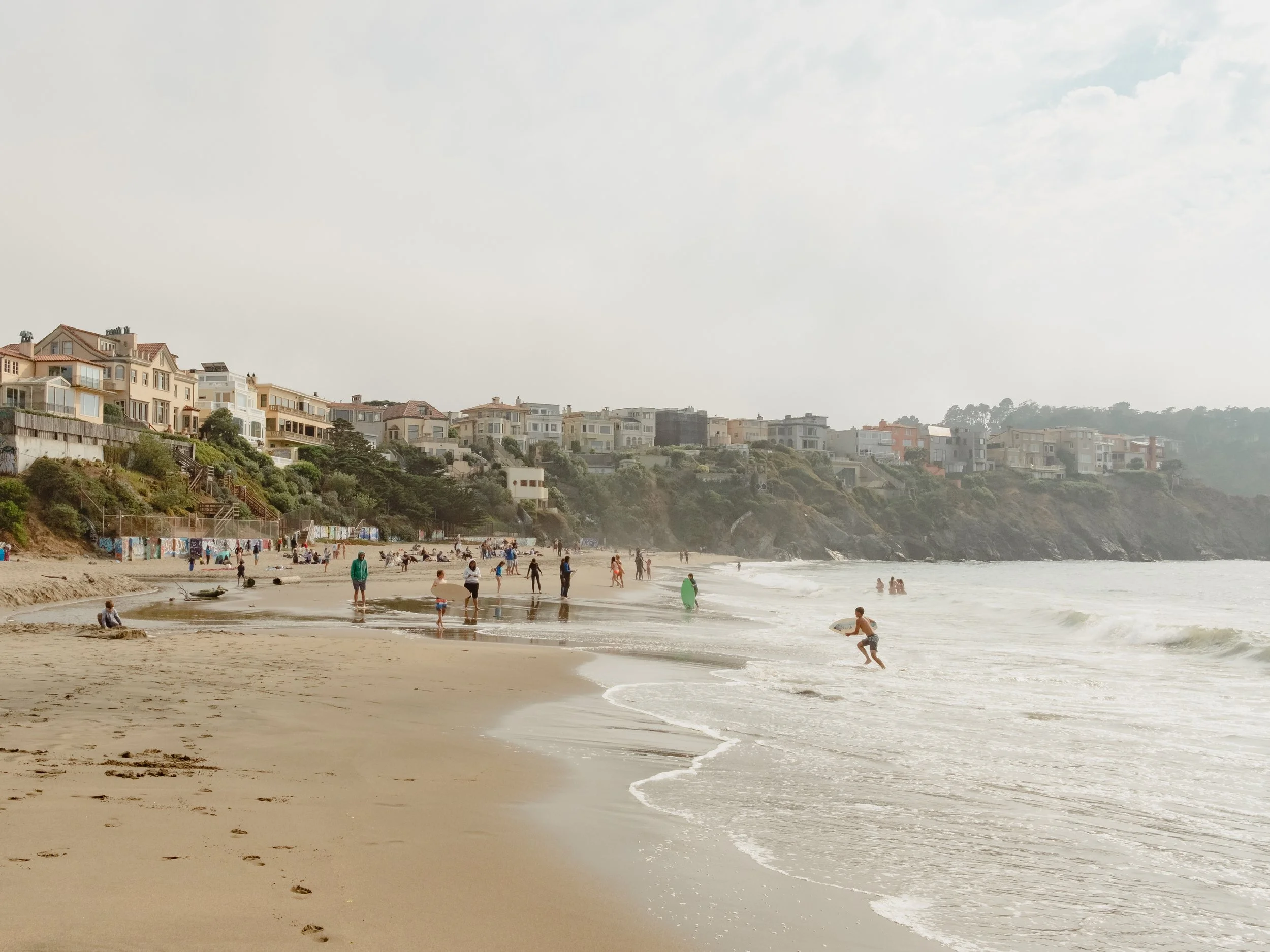People enjoying the beach, walking along the shoreline with houses on a hill in the background on a partly cloudy day.
